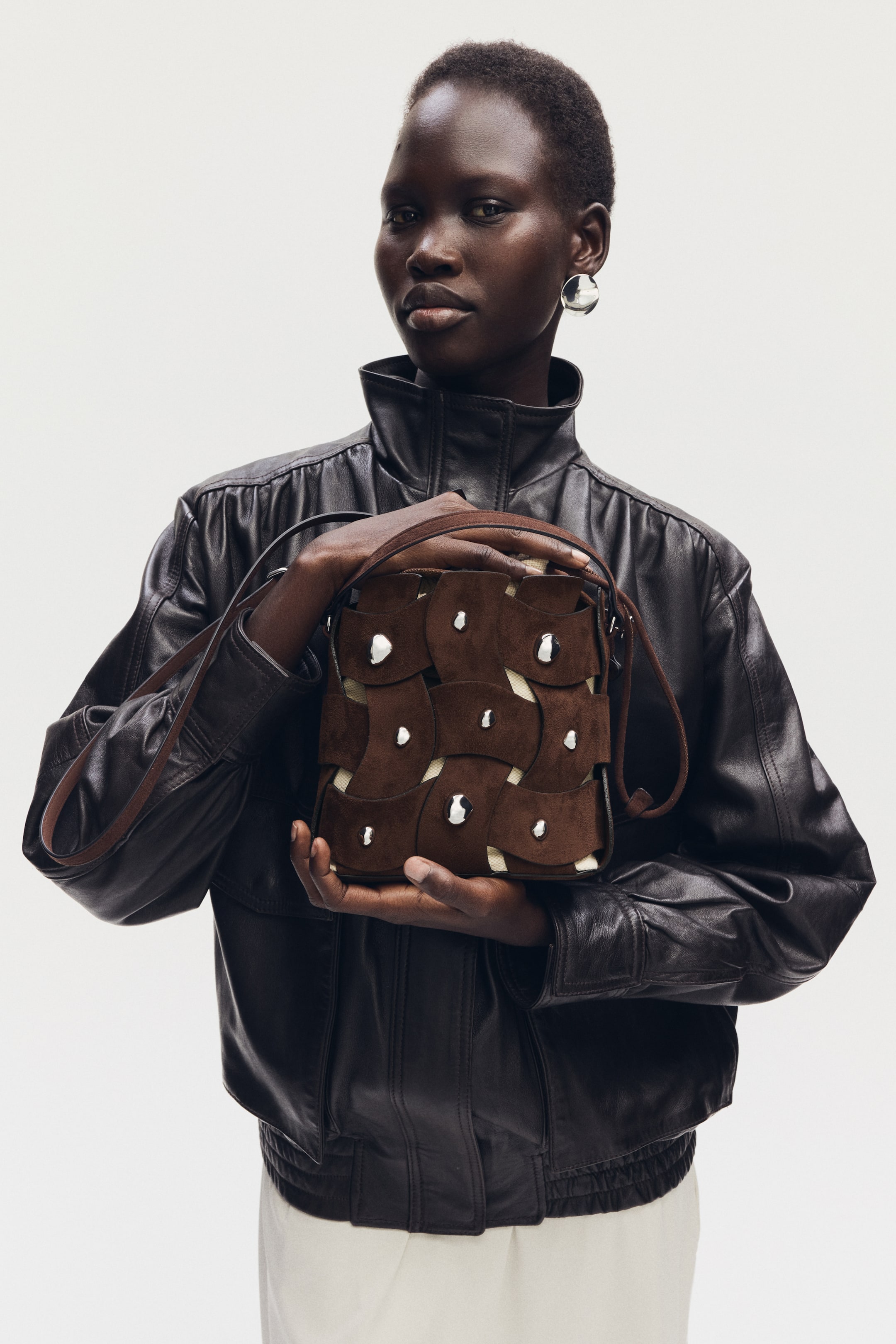 Visualizza immagine più grande: Una borsa marrone viene tenuta frontalmente, mettendo in mostra il suo design con toppe di camoscio marrone scuro disposte in un motivo intrecciato su una base di tessuto più chiaro. Borchie color argento accentuano il design intrecciato e un sottile cinturino marrone è drappeggiato sulla parte superiore.