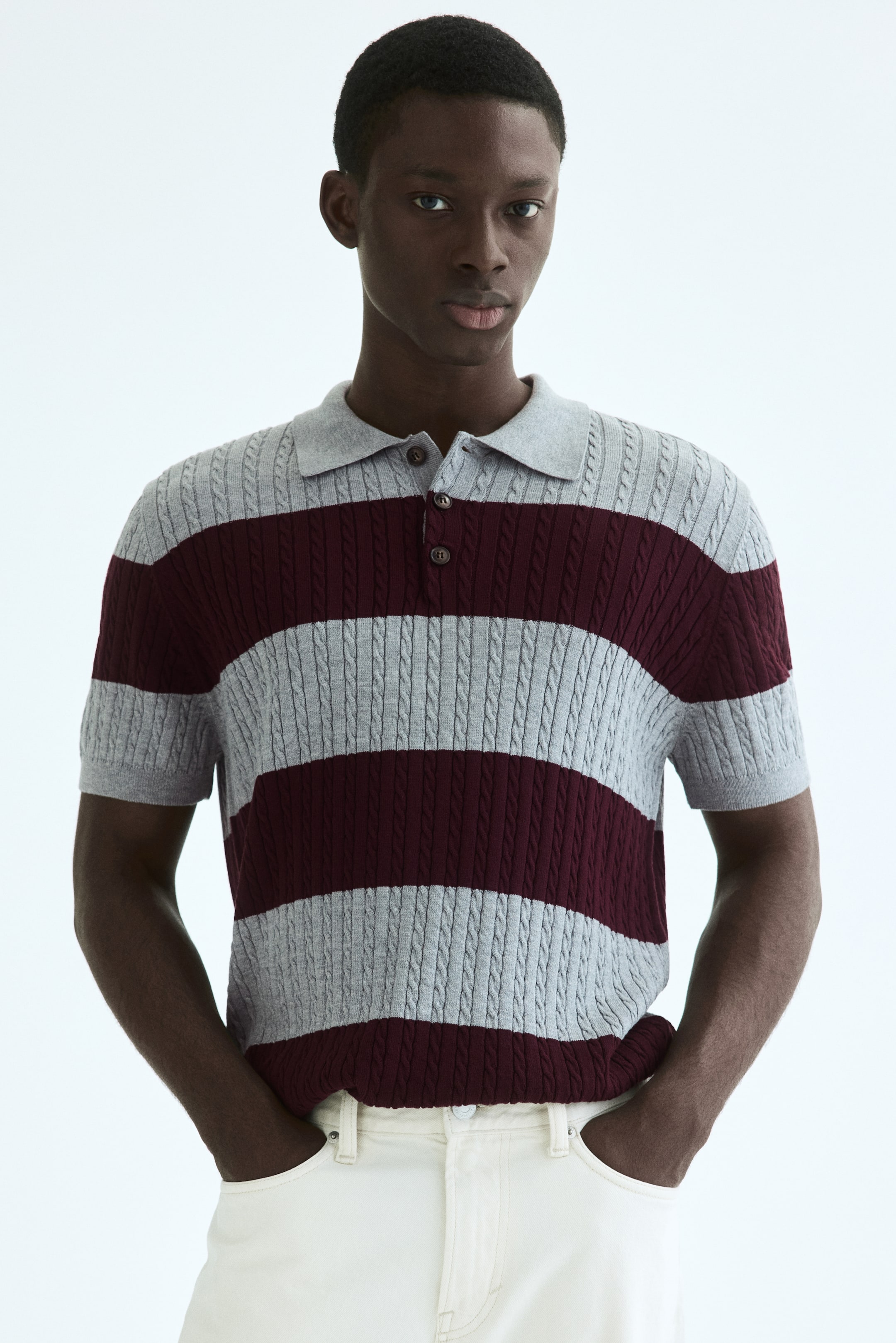 Visualizza immagine più grande: Una maglia polo lavorata a maglia è presentata frontalmente, mostrando strisce orizzontali grigio chiaro e bordeaux con un motivo a trecce. Presenta un colletto polo grigio, un'abbottonatura a tre bottoni con bottoni marrone scuro e maniche corte con polsini a coste.