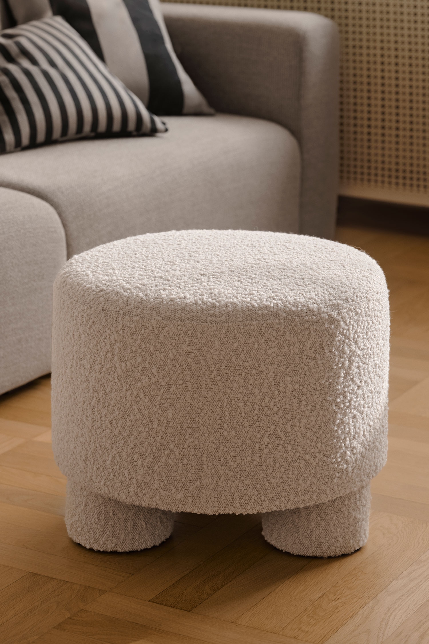 Een ronde poef, bekleed met een gestructureerde bouclÃ© stof in crÃ¨mekleur, staat op drie korte, dikke, bijpassende cilindervormige poten. Het meubelstuk heeft een compact, cilindervormig silhouet met een zacht afgeronde bovenkant en een uniforme bekleding die doorloopt van de zitting tot aan de poten.