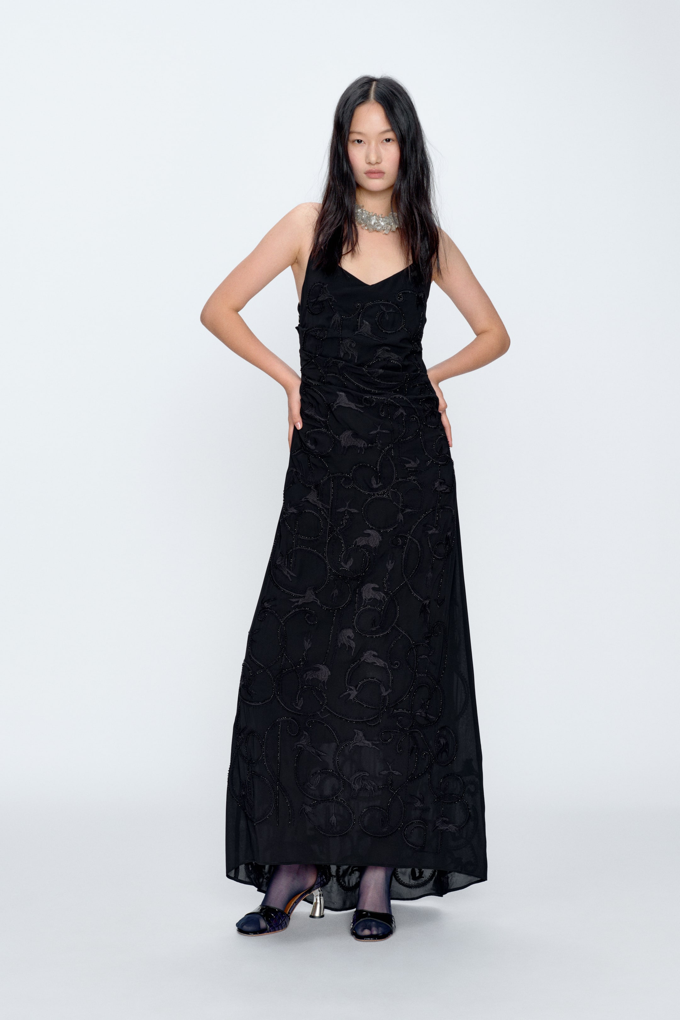 View larger image: A full-length black dress featuring thin spaghetti straps and a V-neckline, with intricate tonal embroidered or beaded swirling patterns across the entire front, from the fitted bodice down to the flowing hem.