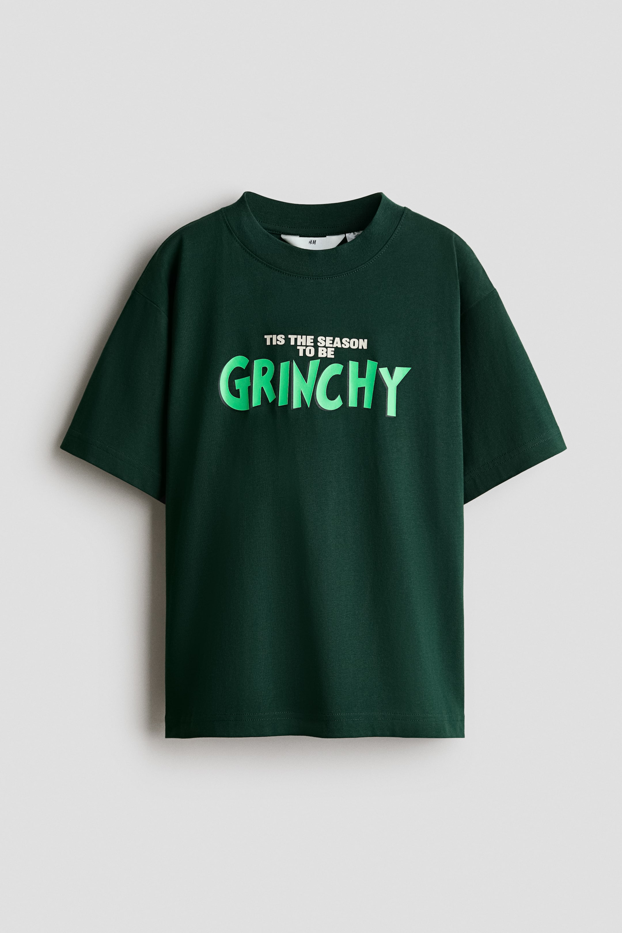 View larger image: A dark green cotton jersey T-shirt is shown from the front, featuring a round, rib-trimmed neckline and dropped shoulders with short sleeves. The chest displays a printed design that reads "TIS THE SEASON TO BE" in small white block letters above "GRINCHY" in large, bright green stylised letters.
