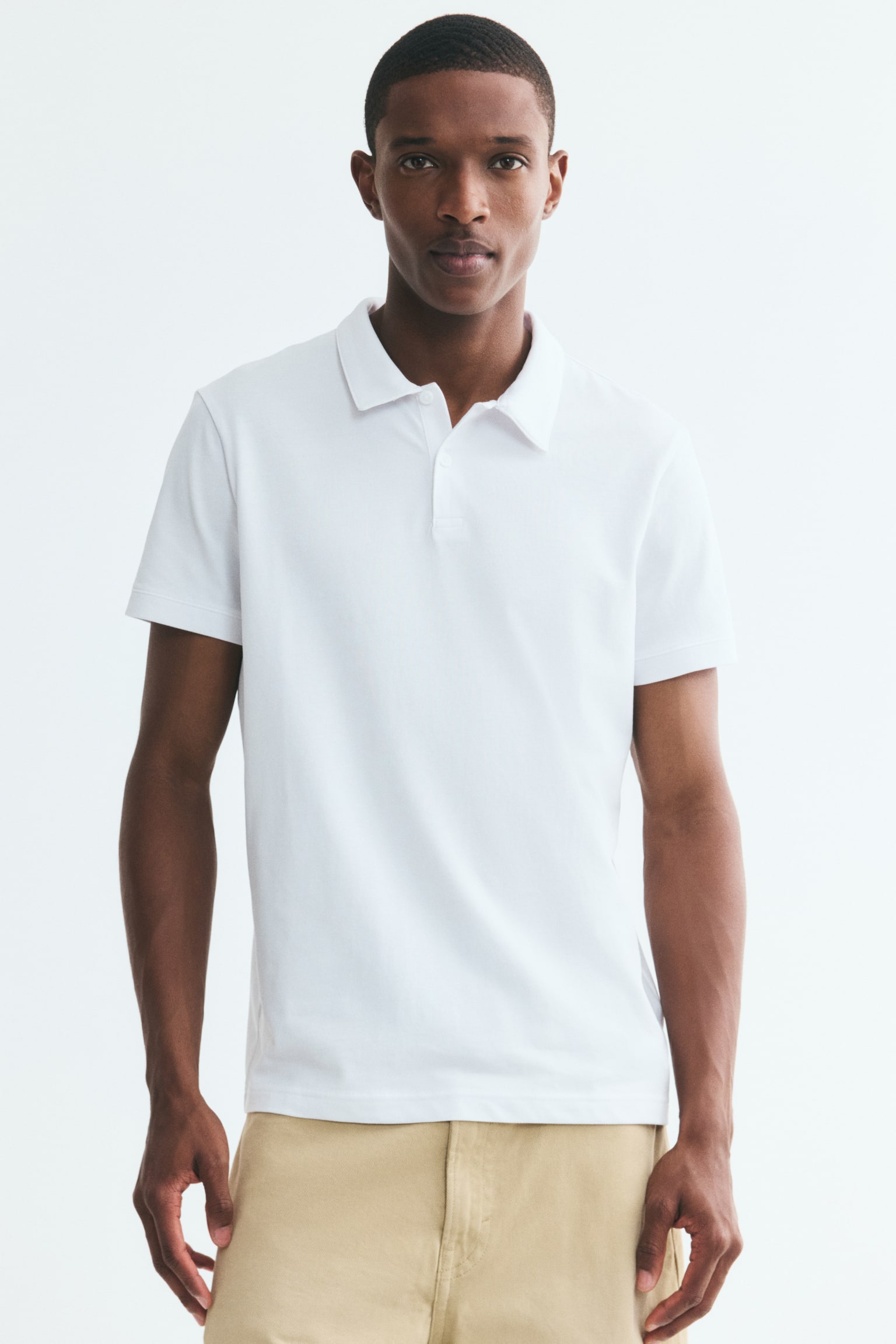 A white polo shirt is displayed from the front, featuring a classic collar and a two-button placket. Short sleeves extend from the shoulders, and a black brand label is visible on the inside of the collar. - 1