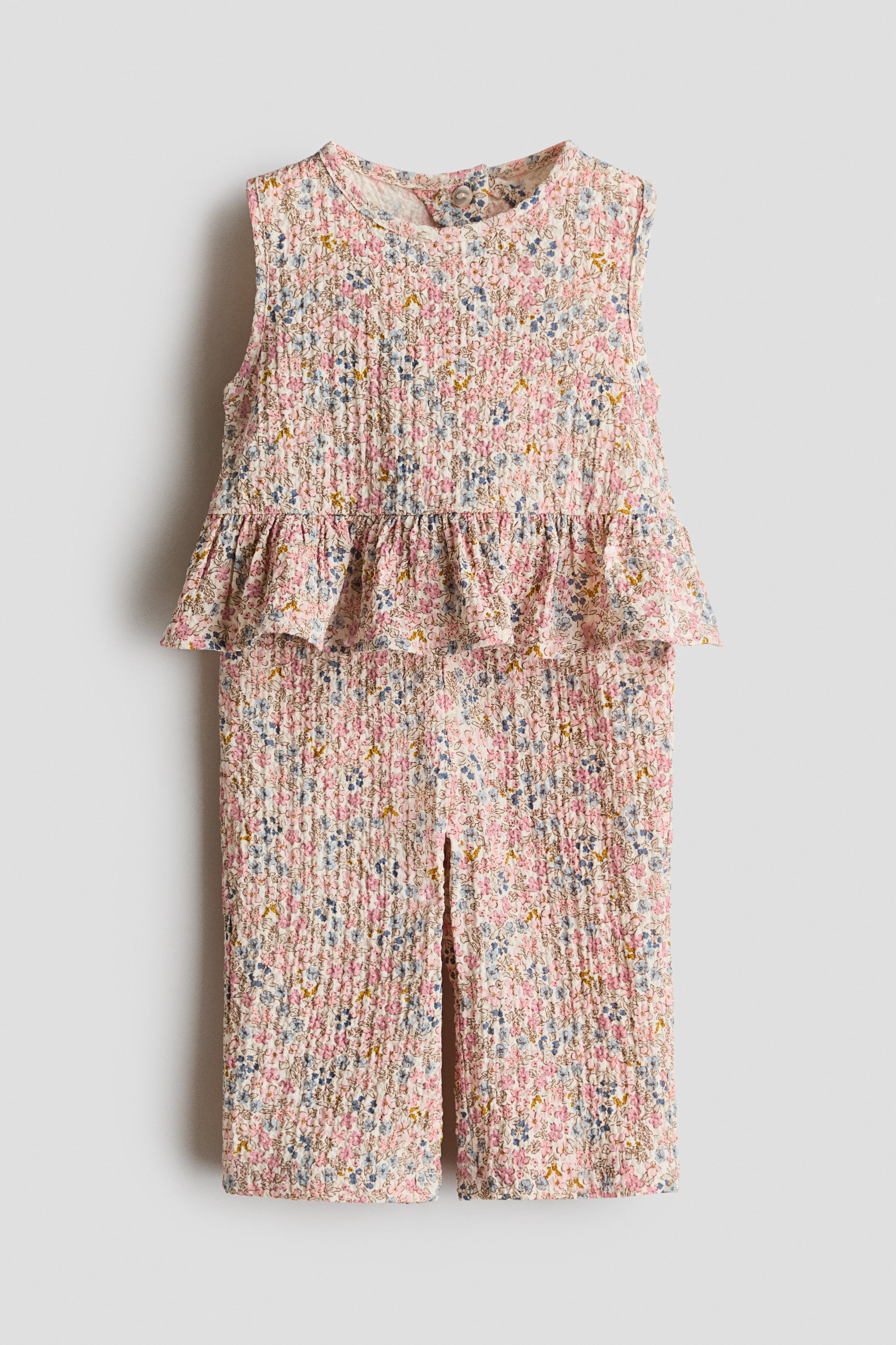 View larger image: A two-piece baby outfit consisting of a sleeveless smocked top with a round neck, peplum hem, and a single button closure at the back, paired with matching smocked pants featuring slits at the ankles. Both pieces showcase a delicate floral print in pink, blue, and yellow on a cream background.
