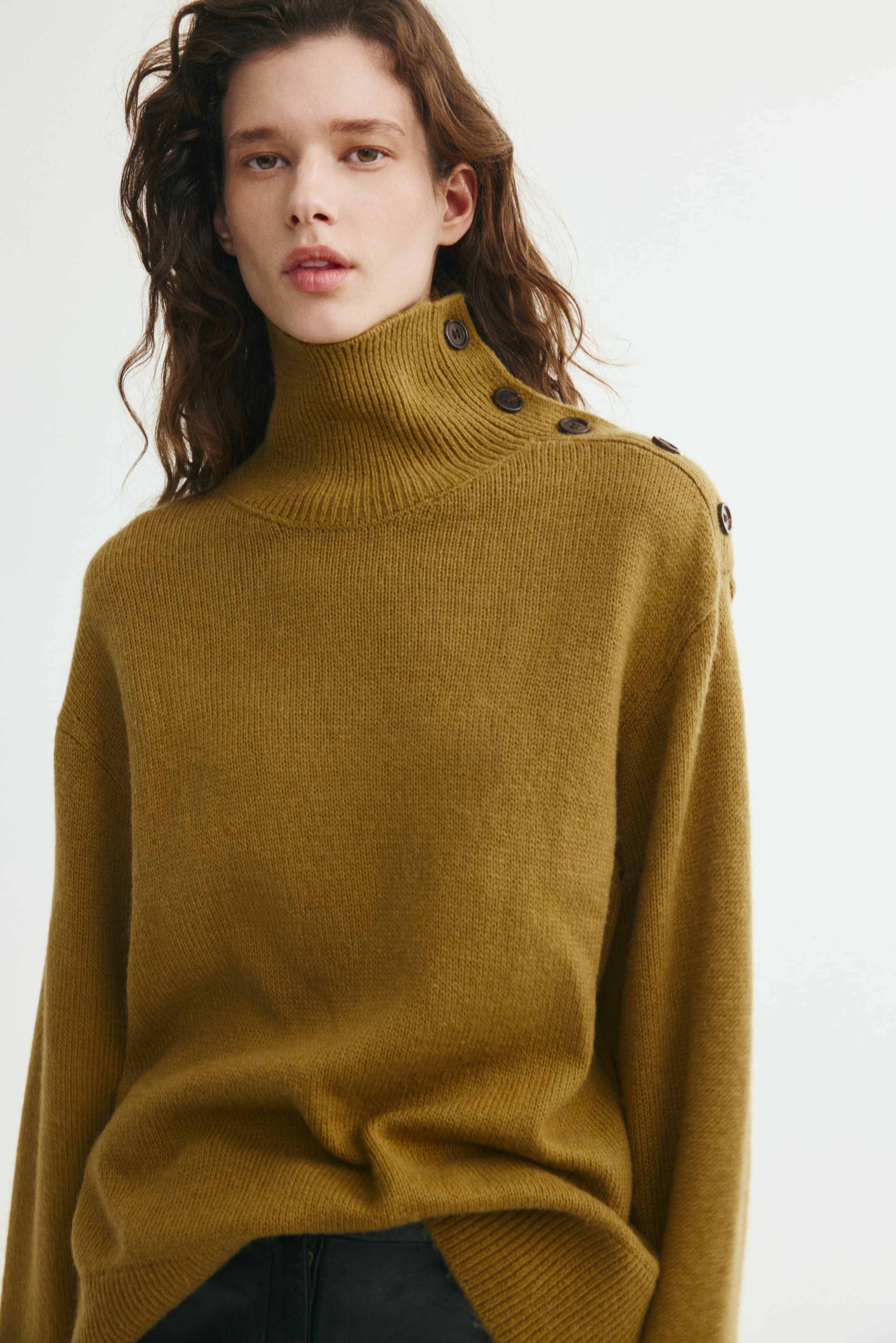 View larger image: A mustard-yellow knit sweater is being worn by the model, featuring a ribbed high neck and a row of decorative dark buttons along the left shoulder. The oversized sweater has dropped shoulders and is shown partially tucked into dark pants at the waist.