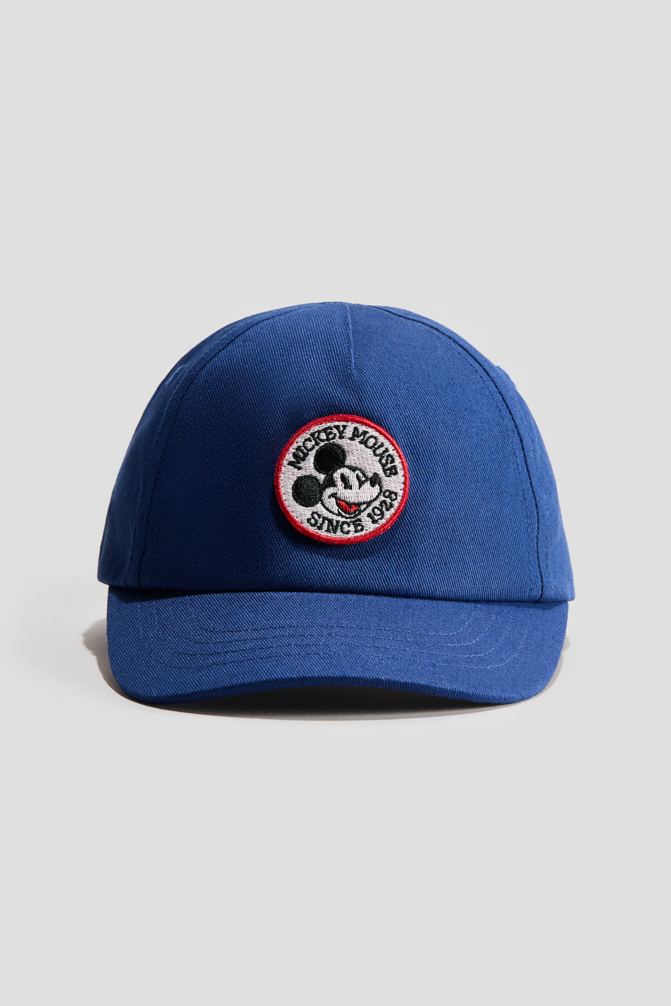 View larger image: A blue cotton twill cap is facing front, showcasing a circular embroidered motif on its front featuring Mickey Mouse's head, the words 'MICKEY MOUSE' and 'SINCE 1928', all outlined by a red border.