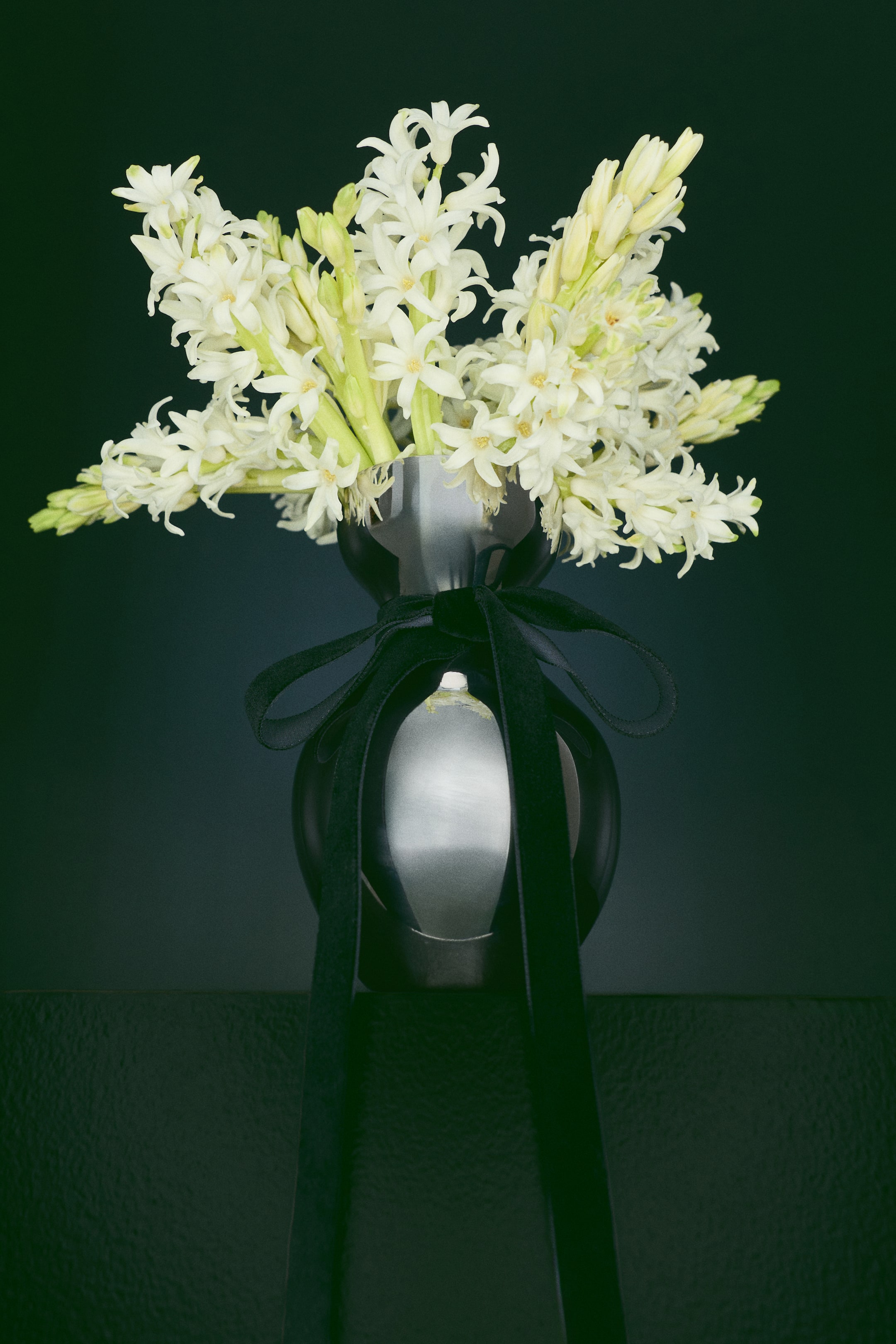 View larger image: A small stainless steel vase with a gourd-style design is shown from the front, holding a full bouquet of white flowers in its wide, flared rim. A black velvet ribbon is tied in a bow around the narrow neck of the reflective metallic body, with long tails draping down the front.