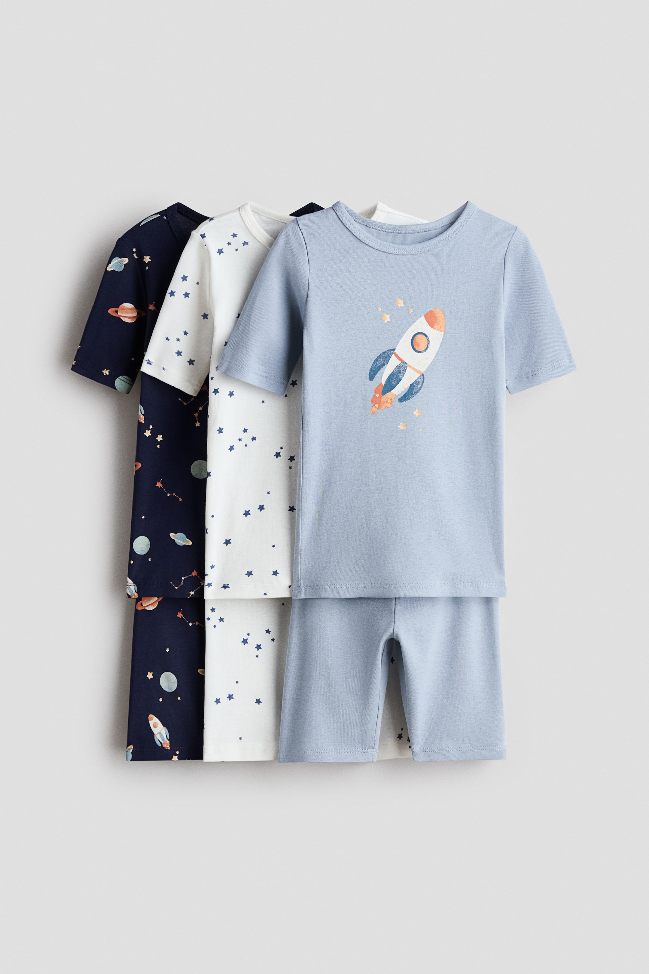 View larger image: The 3-pack Snug Fit Cotton pajamas feature three short-sleeved top and shorts sets, arranged with the front set facing forward. This front set is light blue with a rocket graphic on the top and solid shorts, while the two sets behind it display all-over patterns of blue stars on white and various space elements on dark blue.