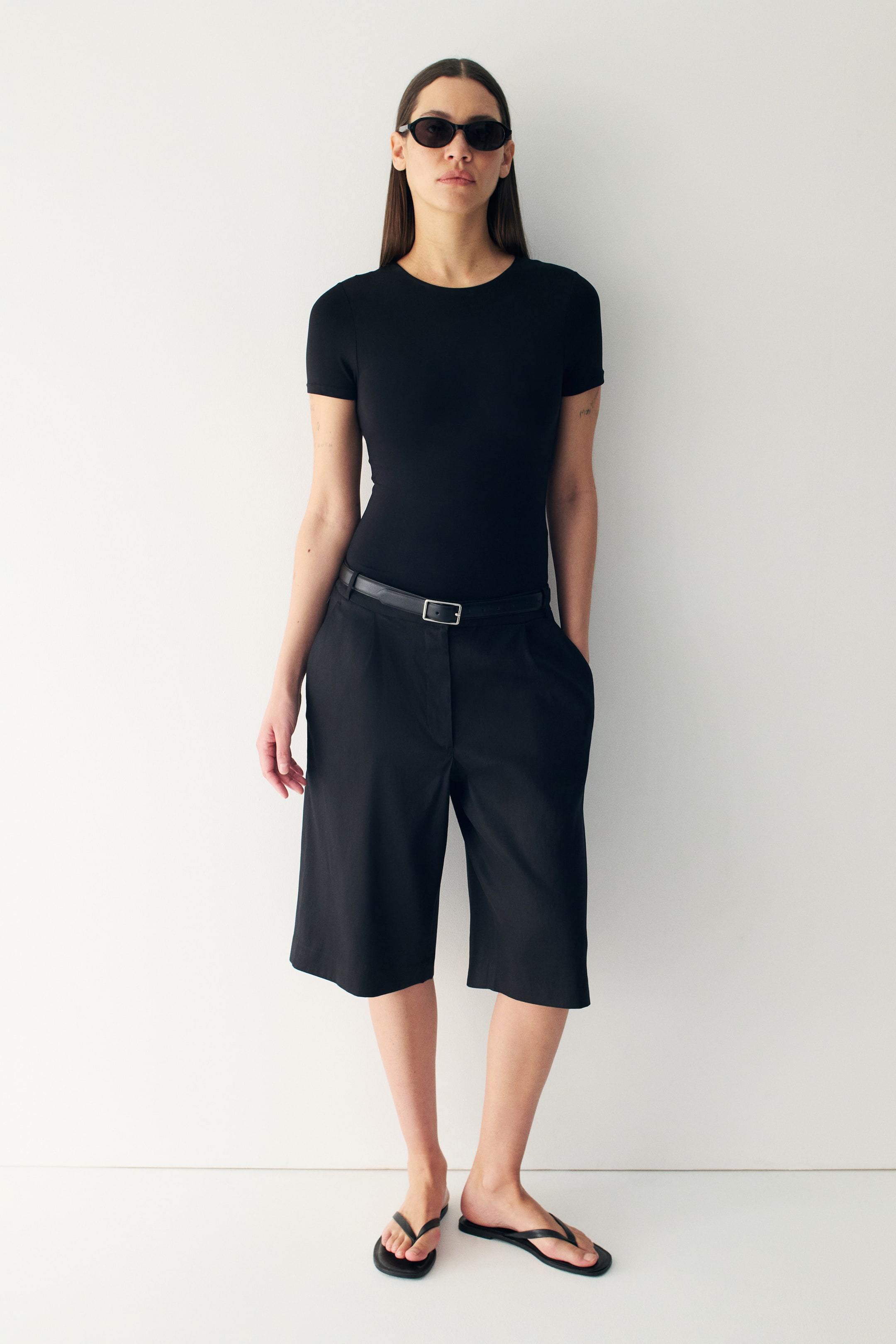 View larger image: A black short-sleeved top fits snugly and is tucked into black, loose-fitting, knee-length shorts. The shorts boast front pleats and pockets, and are cinched with a black belt featuring a silver rectangular buckle.
