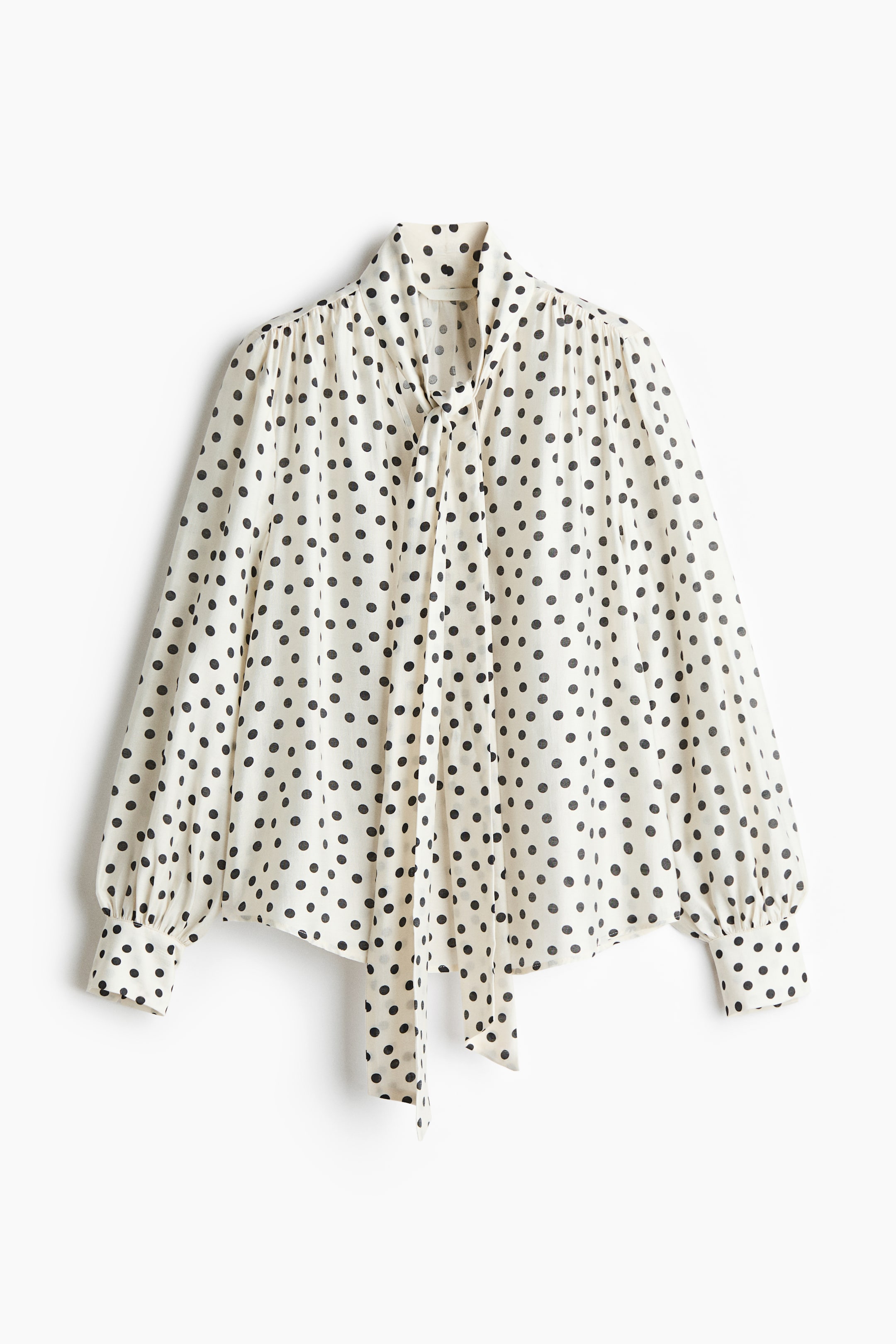 Xem ảnh lớn hơn: Một chiếc áo blouse màu trắng ngà với họa tiết chấm bi đen, nổi bật với cổ đứng và nơ buộc ở cổ, cùng phần tay dài phồng, được nhún ở vai và kết thúc bằng cổ tay cài một nút.