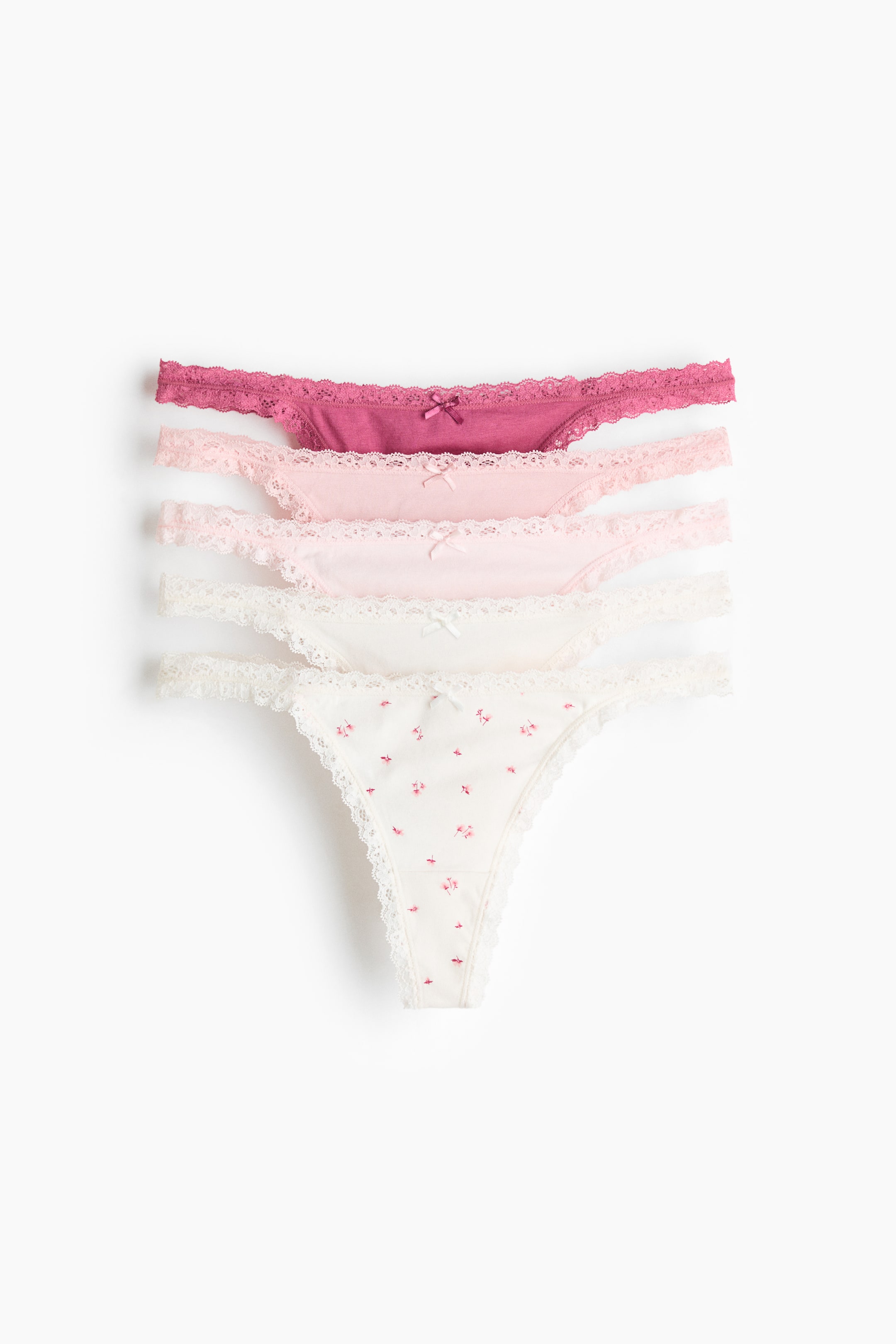 Größeres Bild ansehen: Ein Set aus fünf Baumwoll-Jersey-Stringtangas wird aus der Vorderansicht präsentiert, gestapelt und fächerartig angeordnet. Jeder Stringtanga zeichnet sich durch eine niedrige Taille, schmale Seiten und Spitzenbesätze entlang der Taille und der Beinausschnitte aus, mit einer kleinen Schleife, die die Vorderseite ziert. Die Tangas erscheinen in Nuancen von tiefem Pink, hellem Pink und Off-White, wobei ein Off-White-Tanga ein kleines Blumenmuster aufweist.