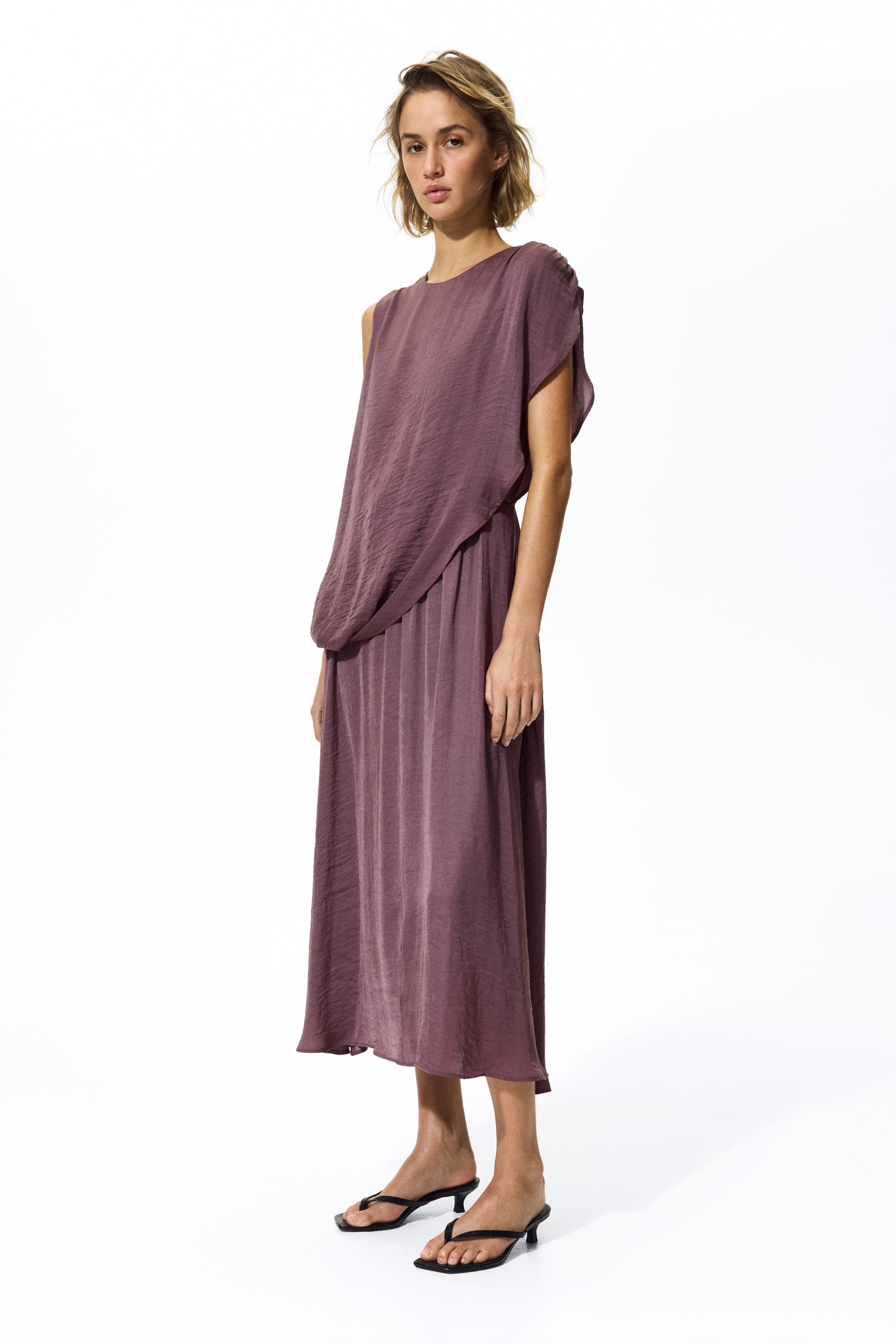 View larger image: A mauve dress showcasing an asymmetrical neckline, with a draped overlay shaping the bodice, and a flowing, midi-length skirt crafted from a subtly crinkled fabric, presented from a side-front angle.