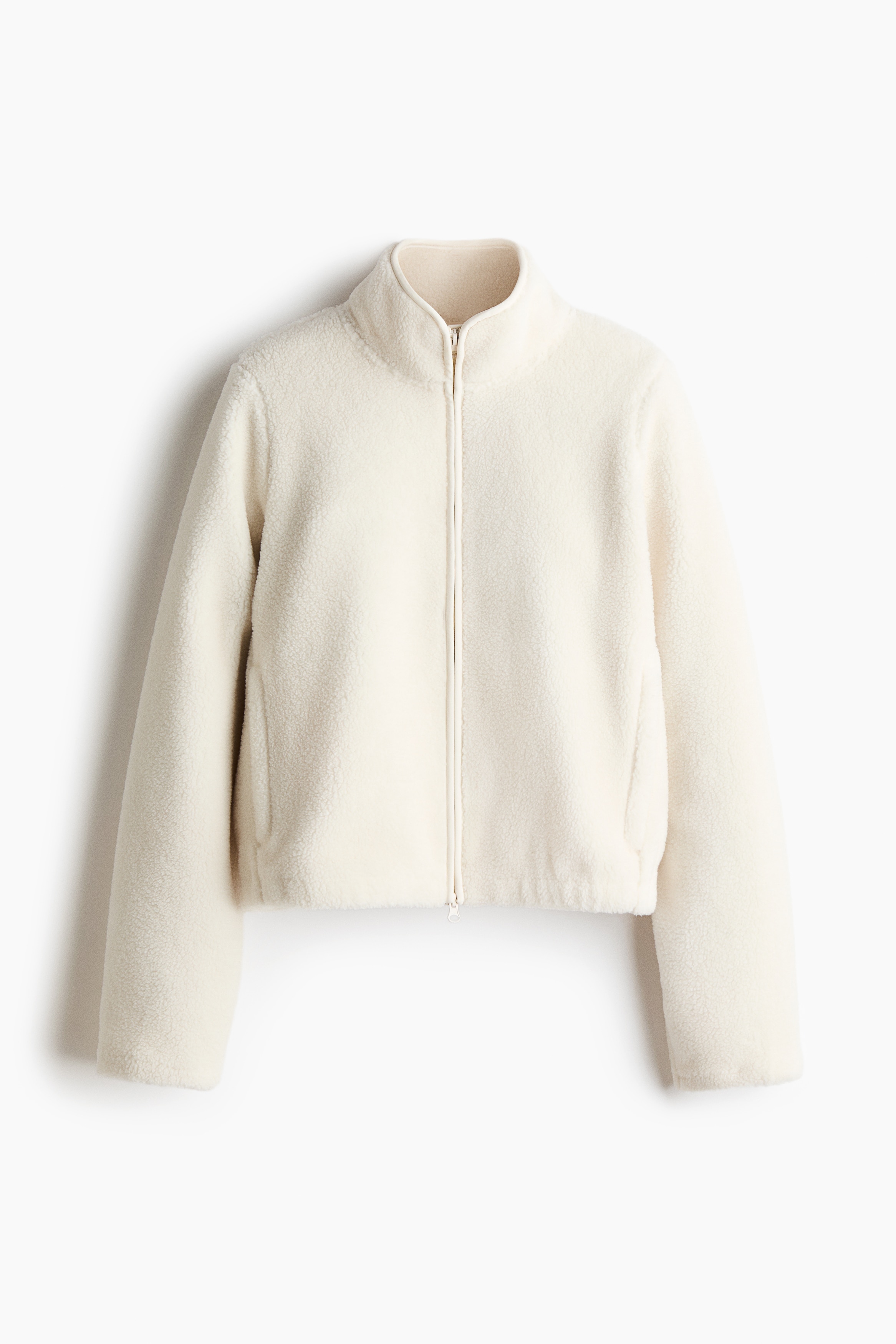 A cream-coloured fleece zip-up jacket with a high collar and open front is being worn by the model. The jacket is layered over a white crop top and paired with light grey drawstring sweatpants and ribbed knit leg warmers.