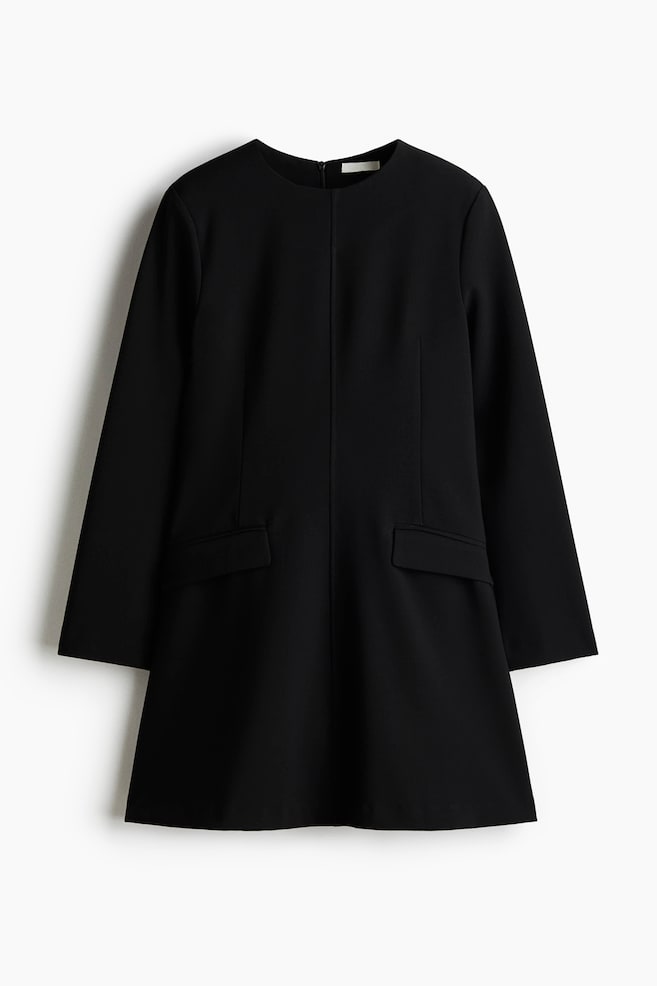 The black short dress is being presented FRONT, featuring a round neckline and long sleeves. Shaping darts are creating a tailored waist, with two flap pockets showing on the front of its flared skirt. - 1