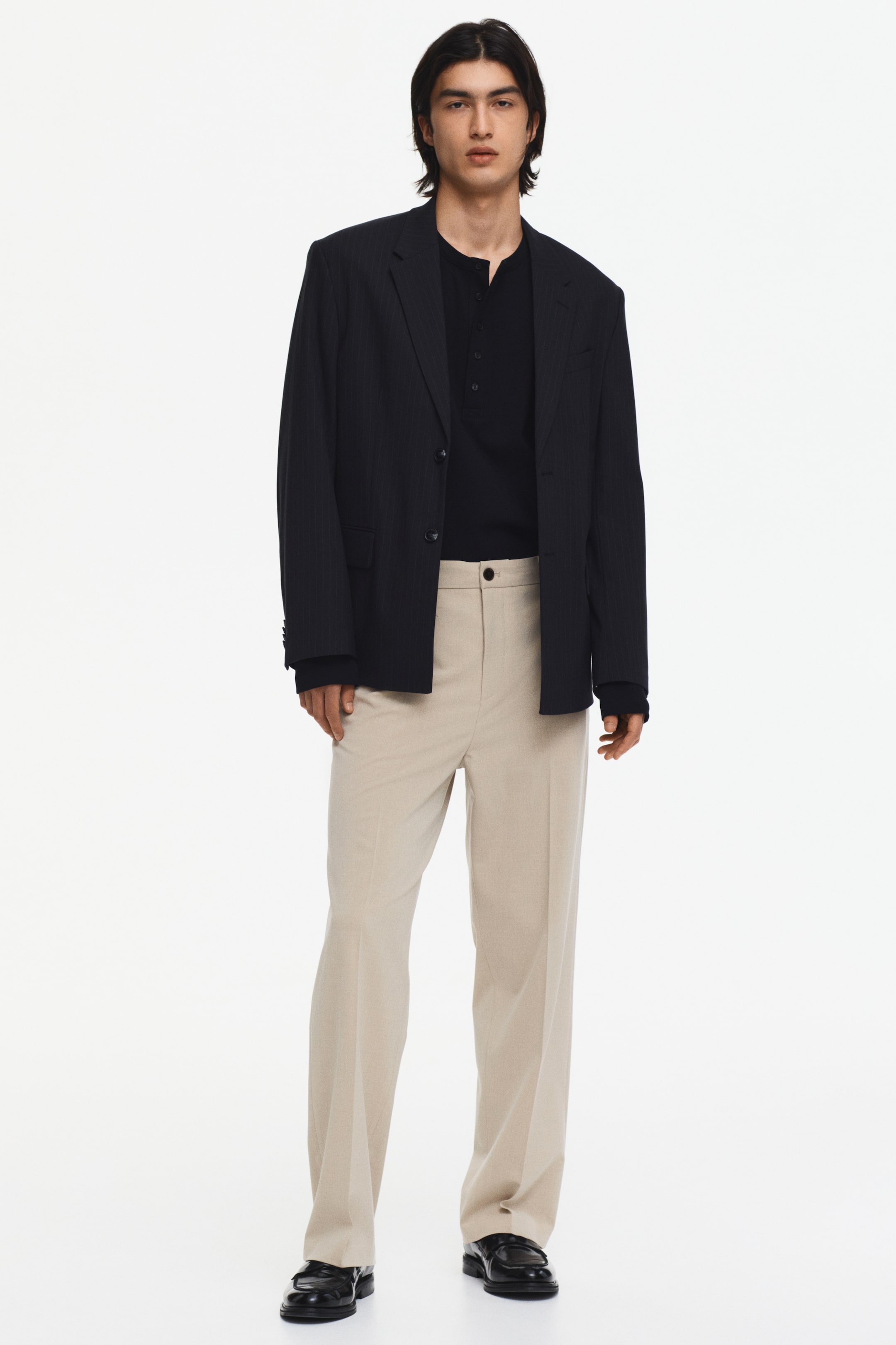 View larger image: A front-facing full outfit is showcased, featuring a dark blue pinstriped blazer with notch lapels and a two-button closure, layered over a black long-sleeve Henley shirt. Light beige wide-leg trousers with a flat front and a single visible button are worn, styled with black penny loafers.