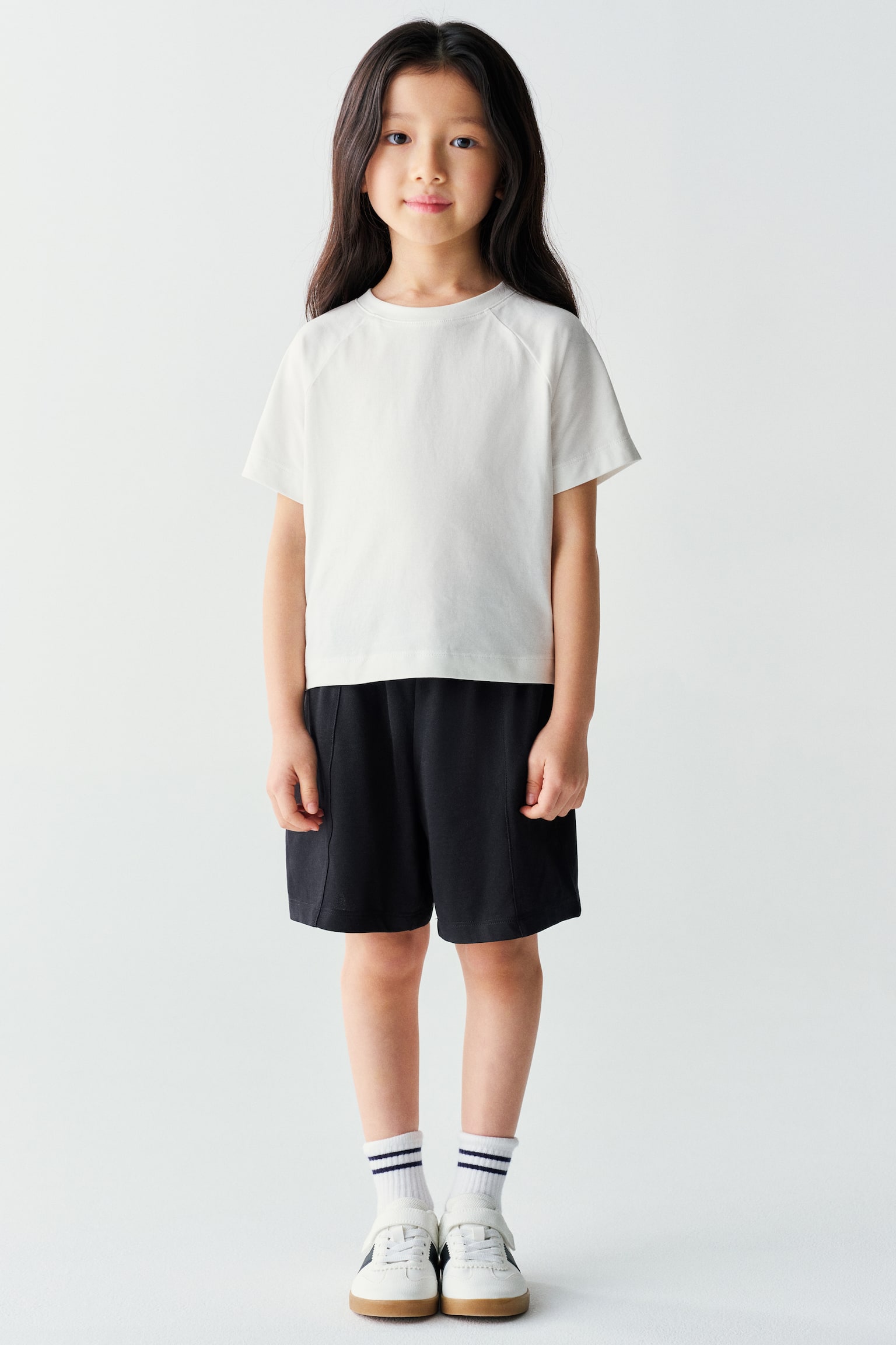 The model is wearing a white, short-sleeved raglan T-shirt and black mid-length shorts. The casual outfit is styled with striped white crew socks and white sneakers. - 1