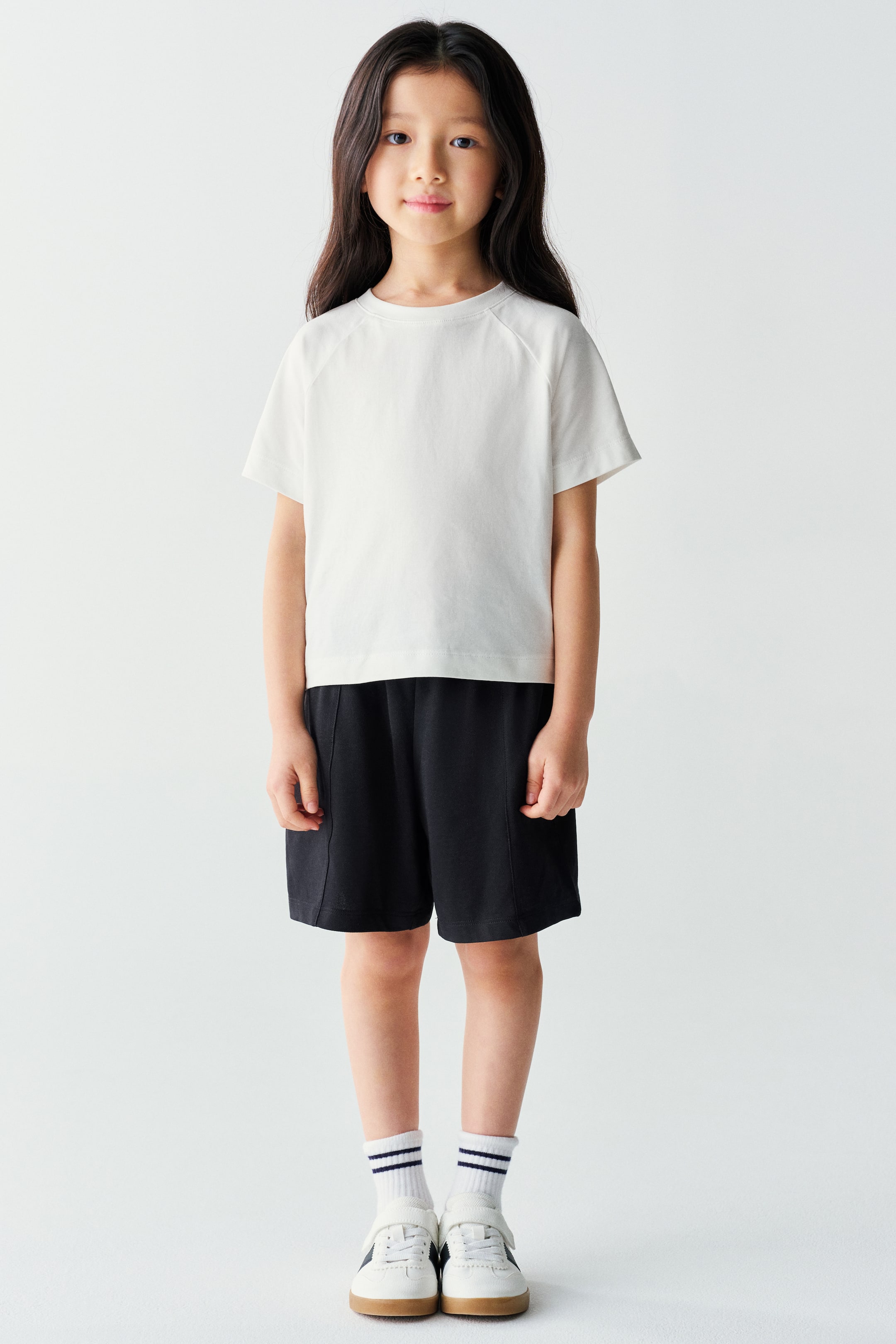 Xem ảnh lớn hơn: Người mẫu đang mặc một chiếc áo thun raglan tay ngắn màu trắng và quần short lửng màu đen. Bộ trang phục thường ngày được phối cùng tất cổ ngắn màu trắng kẻ sọc và giày sneaker trắng.