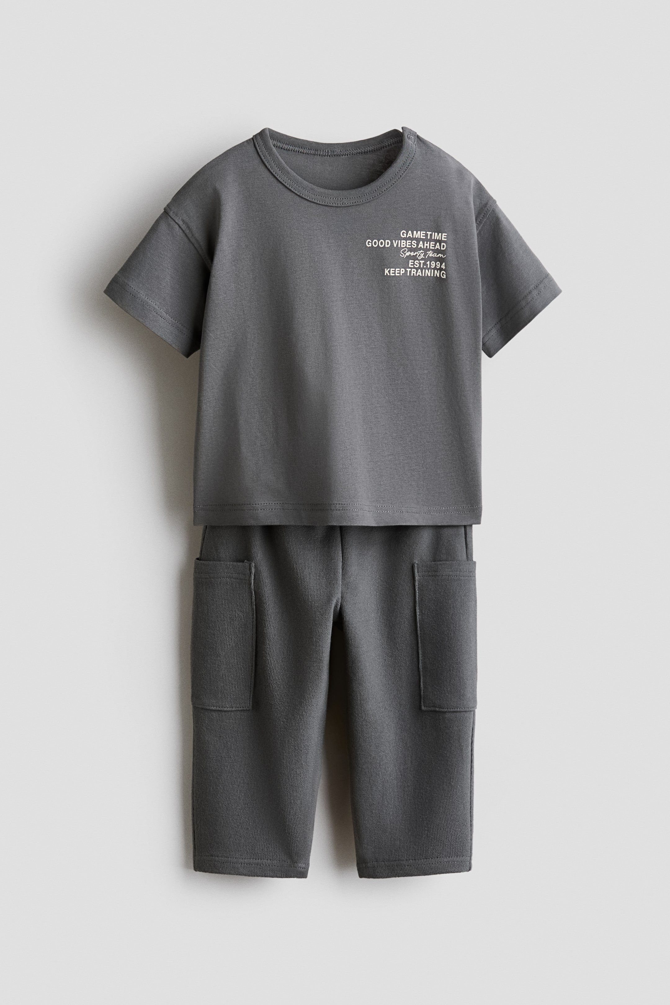 View larger image: A dark grey two-piece outfit displays a short-sleeved T-shirt and matching pants, shown from the front. The T-shirt features white text on the chest, and the pants have two cargo pockets on the sides, both made from a subtly textured fabric.