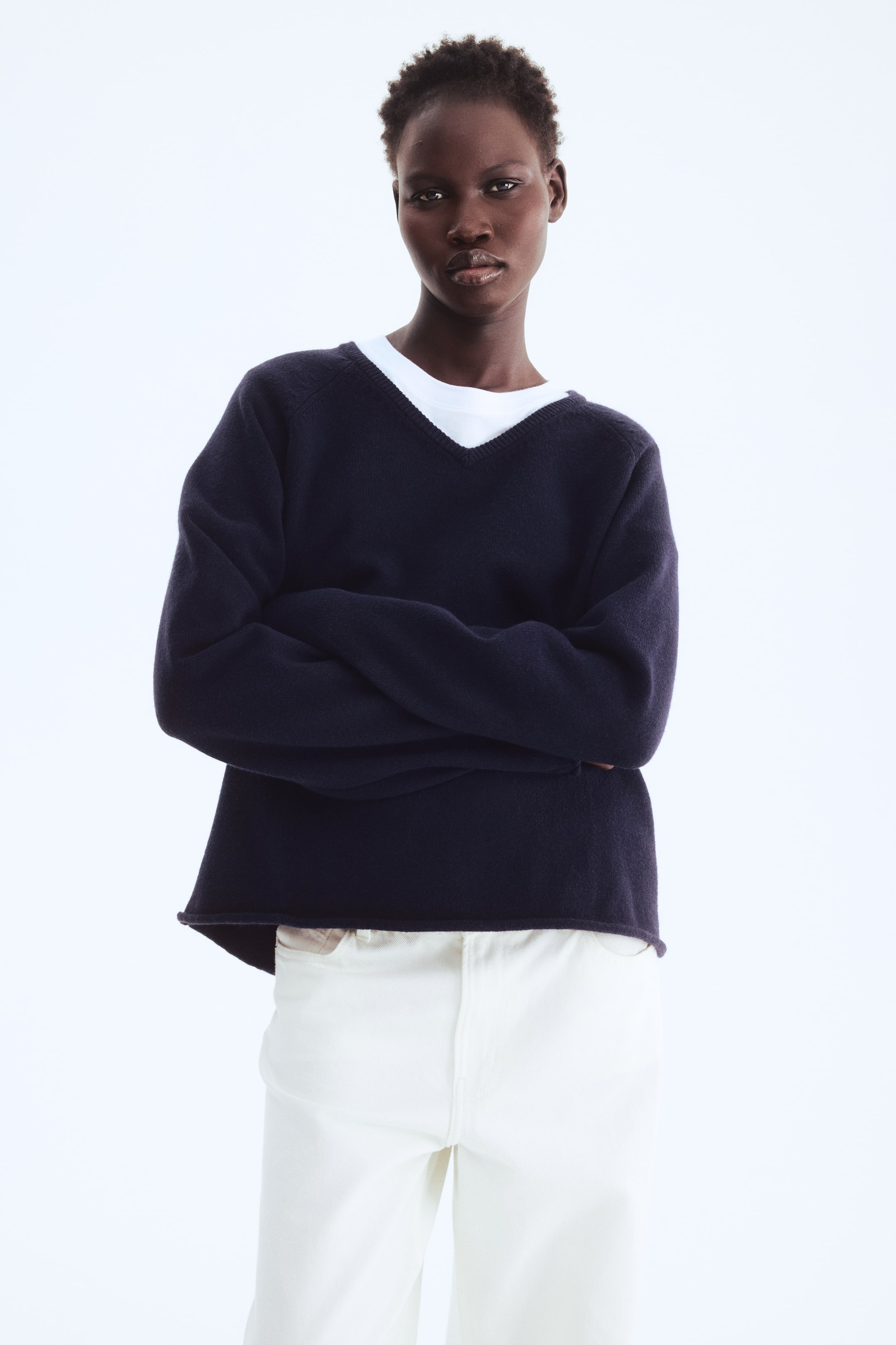 View larger image: The model is wearing an oversized dark blue jumper featuring a ribbed V-neck, long raglan sleeves, and ribbed roll edges at the cuffs and hem, with arms crossed. Underneath the V-neck, a white t-shirt is layered, complemented by light-coloured trousers.