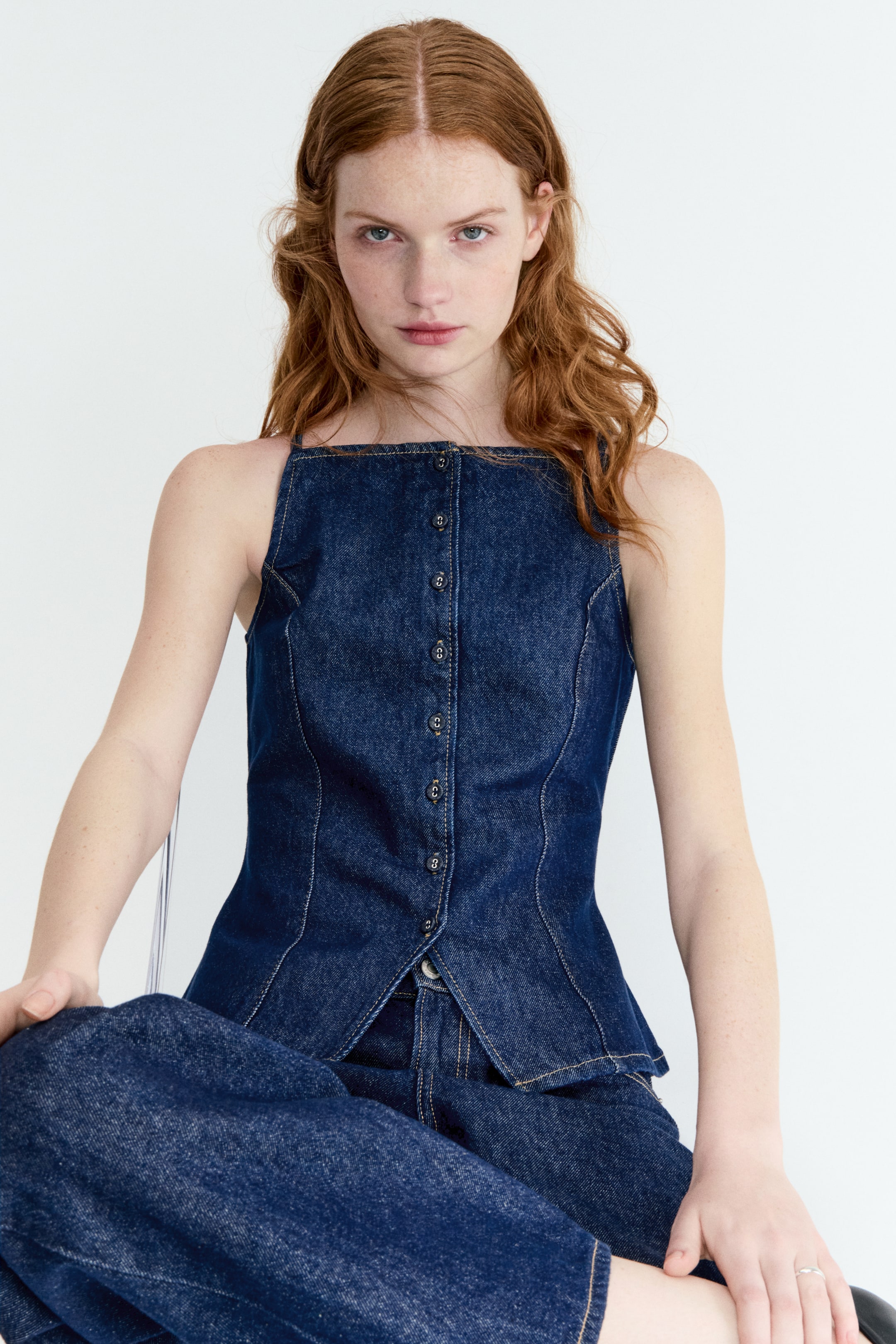 Ver imagen más grande: Un top de mezclilla azul oscuro se presenta de frente, con un escote cuadrado y tirantes finos. Está diseñado con un cierre completo de botones pequeños y oscuros en la parte delantera, costuras de contraste visibles y una silueta ajustada que termina en un dobladillo delantero en punta.