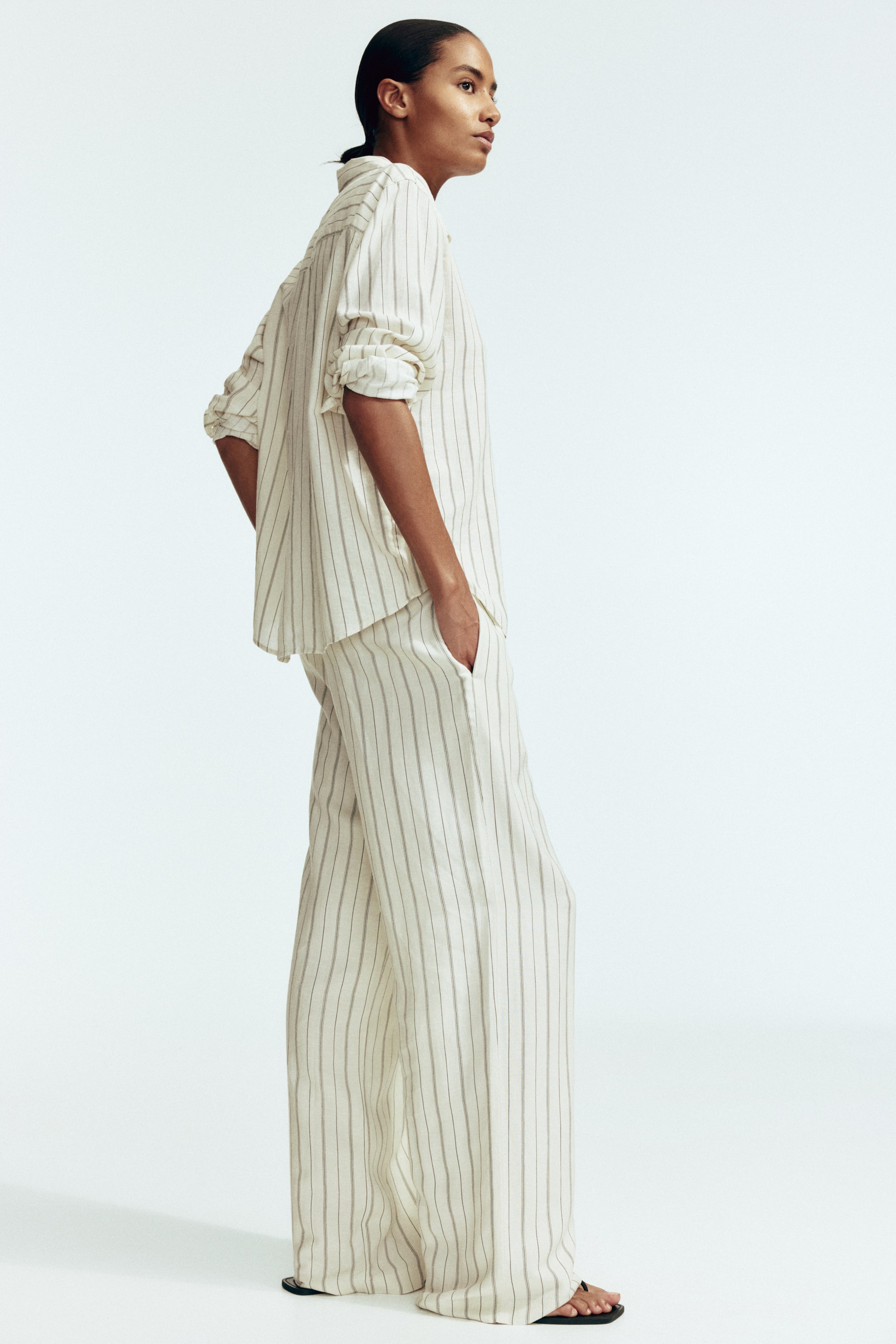 View larger image: A cream pinstripe button-up shirt and matching wide-leg pants are shown in a side-profile view. The shirt features rolled-up sleeves, and the relaxed-fit pants show a hand resting in one of its pockets.