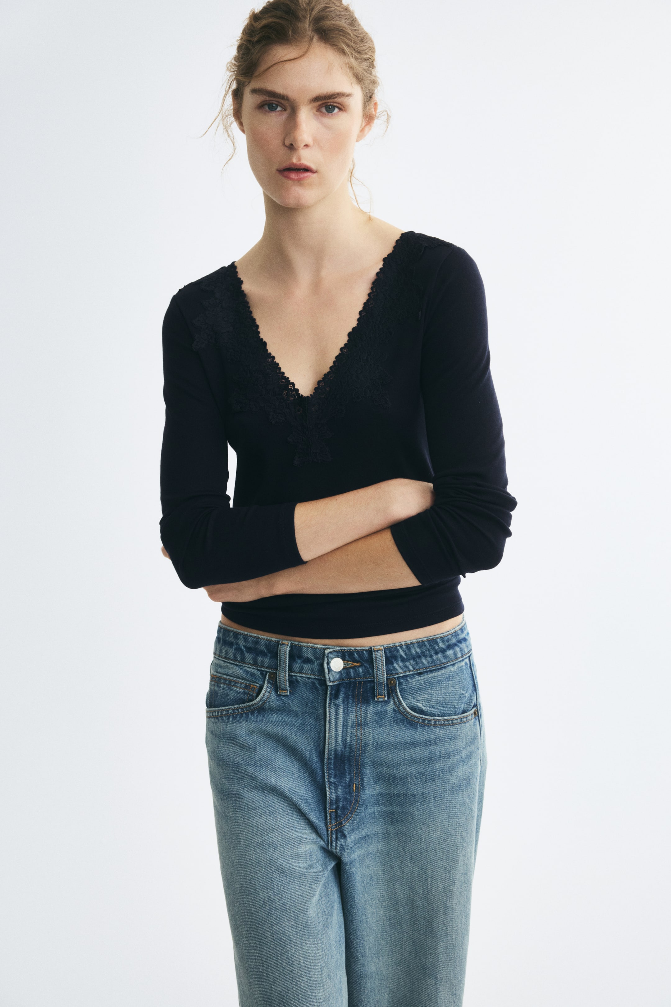 View larger image: A dark blue, fitted rib-knit top featuring a lace-trimmed V-neckline and long sleeves is worn by the model, who has her arms crossed. The top is styled with a pair of light-wash blue jeans.