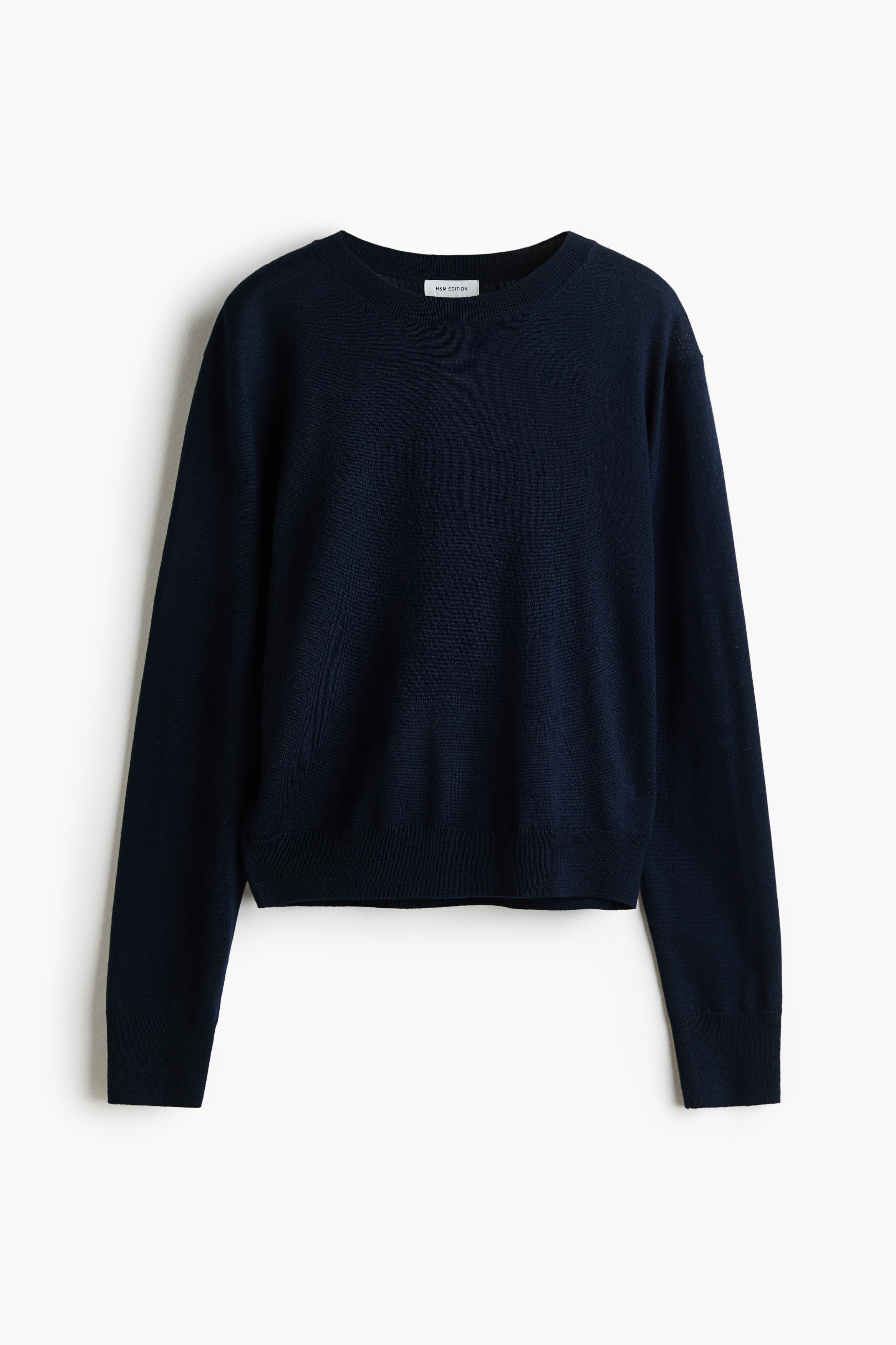 The model is wearing a navy blue knitted wool jumper featuring a round neckline, dropped shoulders, and long sleeves with ribbed cuffs. The jumper is styled tucked into blue jeans with a black belt.