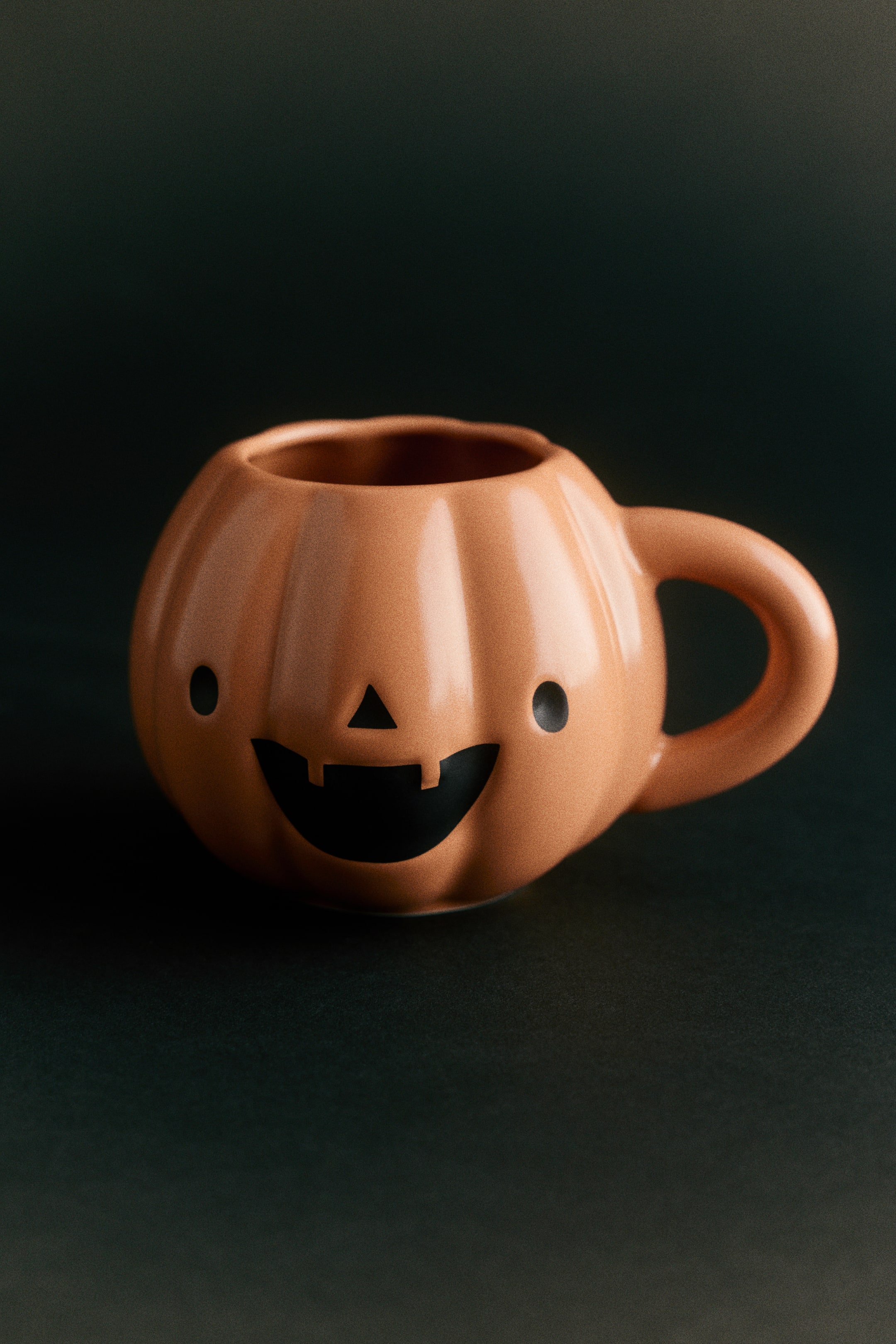 Agrandir l'image: Une tasse en porcelaine en forme de citrouille est orientée vers l’avant, avec un corps côtelé orange clair et une anse épaisse incurvée sur la droite. L’avant de la tasse arbore un visage de jack-o’-lantern minimaliste noir, composé de deux yeux ovales, d’un petit nez triangulaire et d’une large bouche souriante.