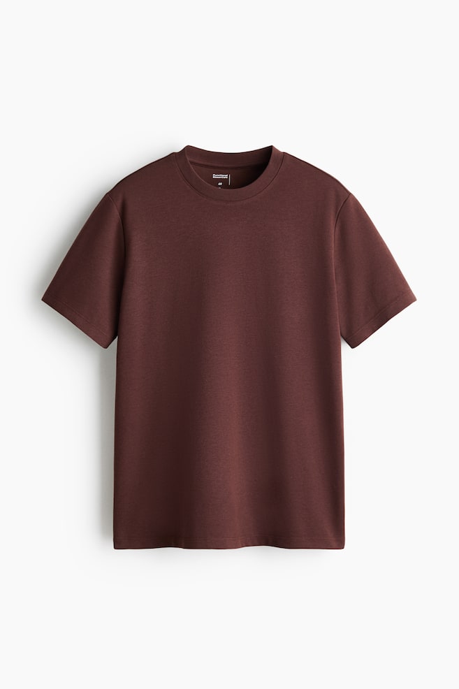 Una T-shirt marrone scuro è mostrata dal davanti, caratterizzata da uno scollo rotondo, maniche corte e un orlo a taglio dritto. Un'etichetta bianca è visibile sul colletto interno posteriore. - 1