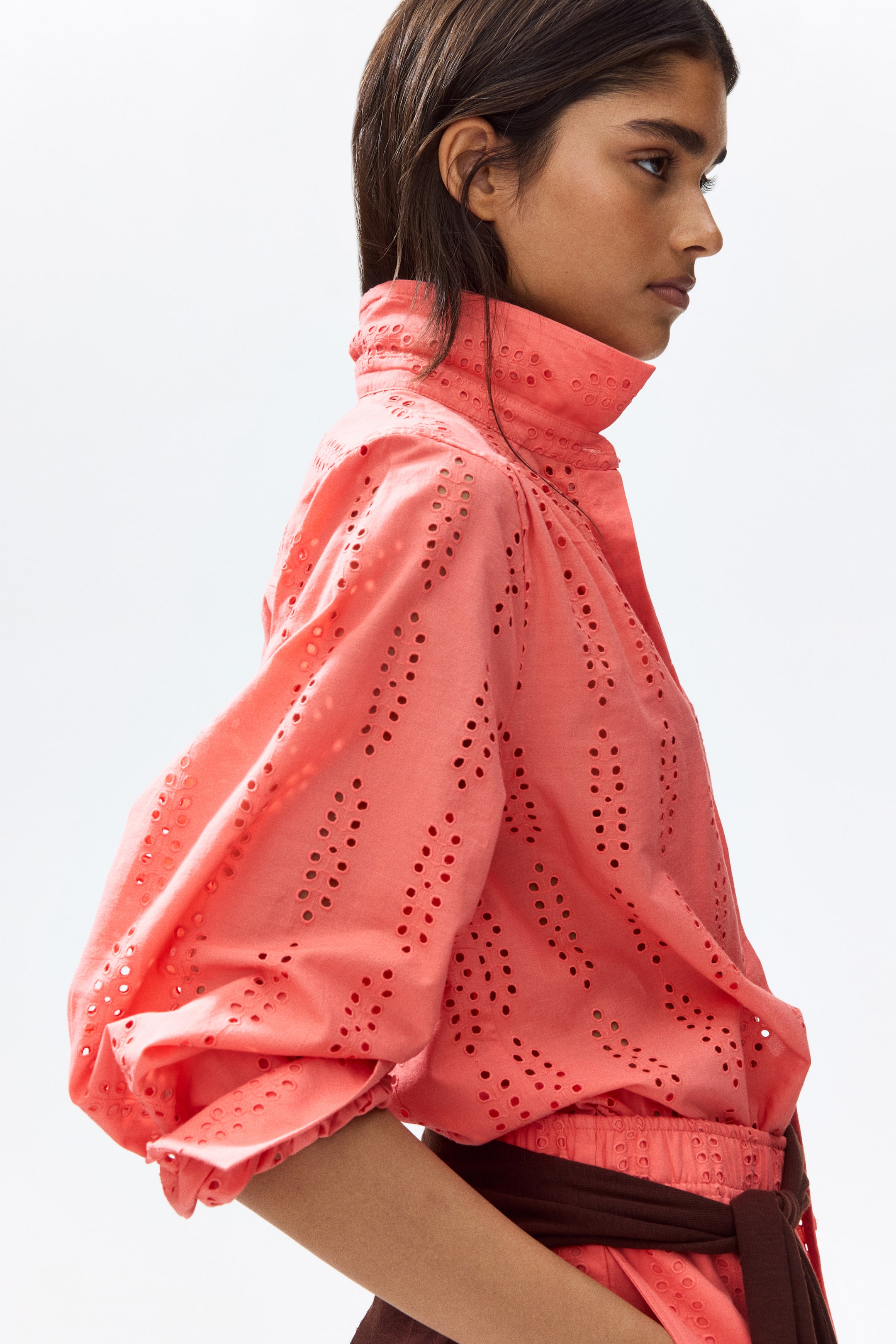 View larger image: The model is showcasing a coral relaxed-fit shirt adorned with broderie anglaise. It boasts a standing collar with a visible button, a gathered yoke, and long balloon sleeves. The waist is elegantly cinched by a dark brown sash.