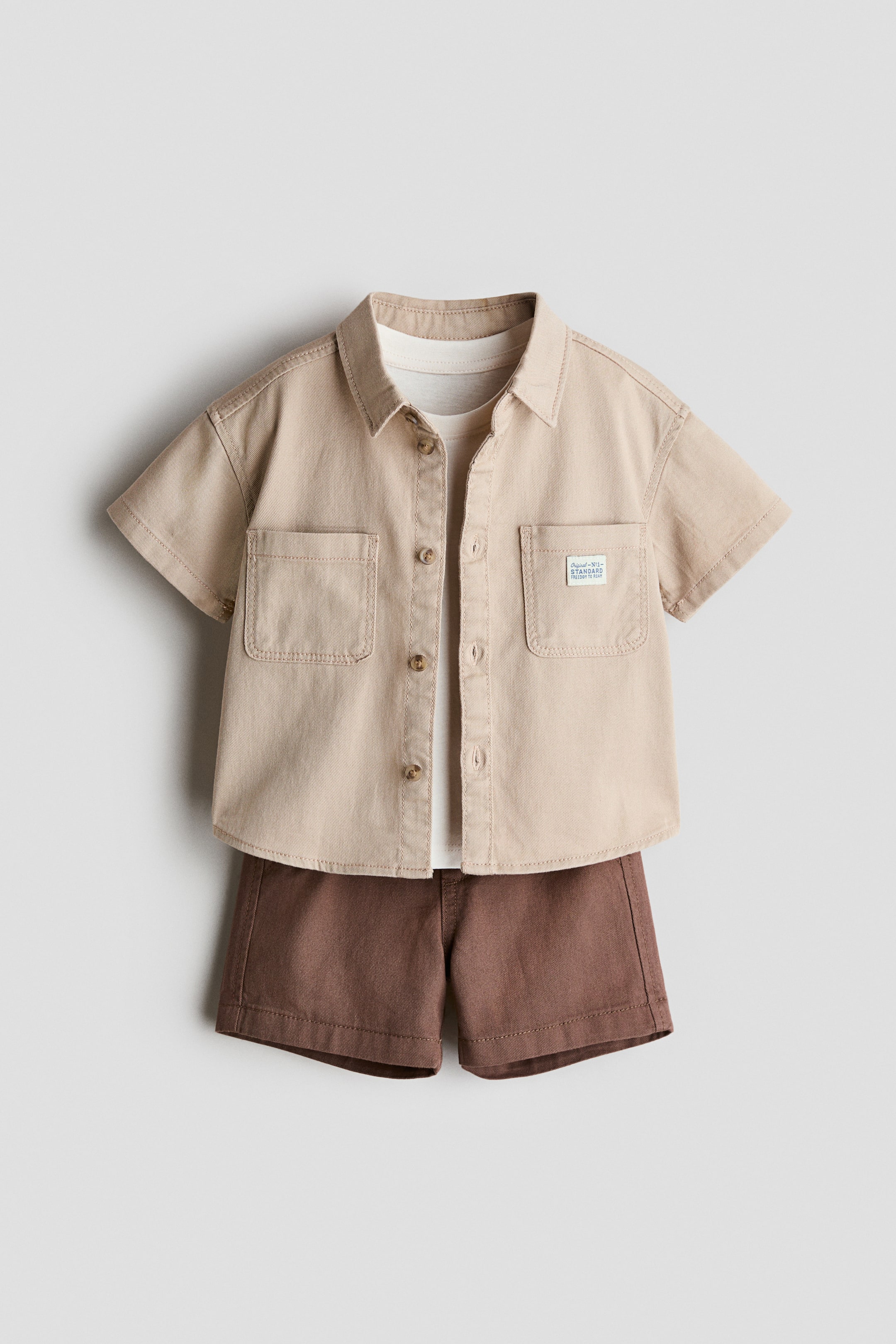 View larger image: The 3-piece cotton ensemble showcases a beige short-sleeved button-down shirt with a pointed collar and two patch pockets on the chest, one adorned with a small label. It's styled open over a white crew-neck t-shirt and paired with brown shorts.