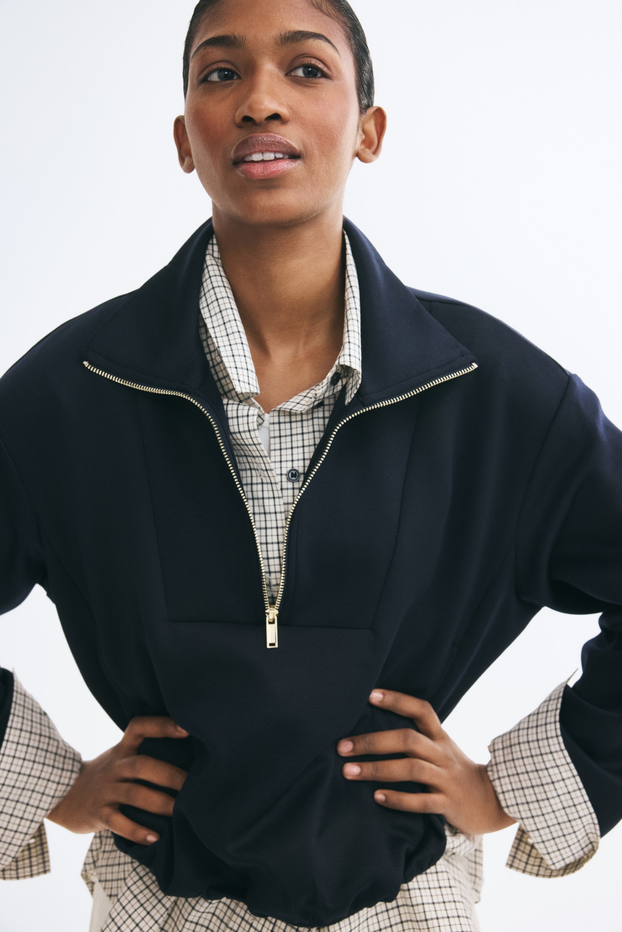 View larger image: A navy blue scuba-fabric sweater with a stand collar and gold half-zip closure is worn by the model over a grid-patterned shirt. The model is posing with hands on the hips, highlighting the sweater’s dropped shoulders and relaxed silhouette.