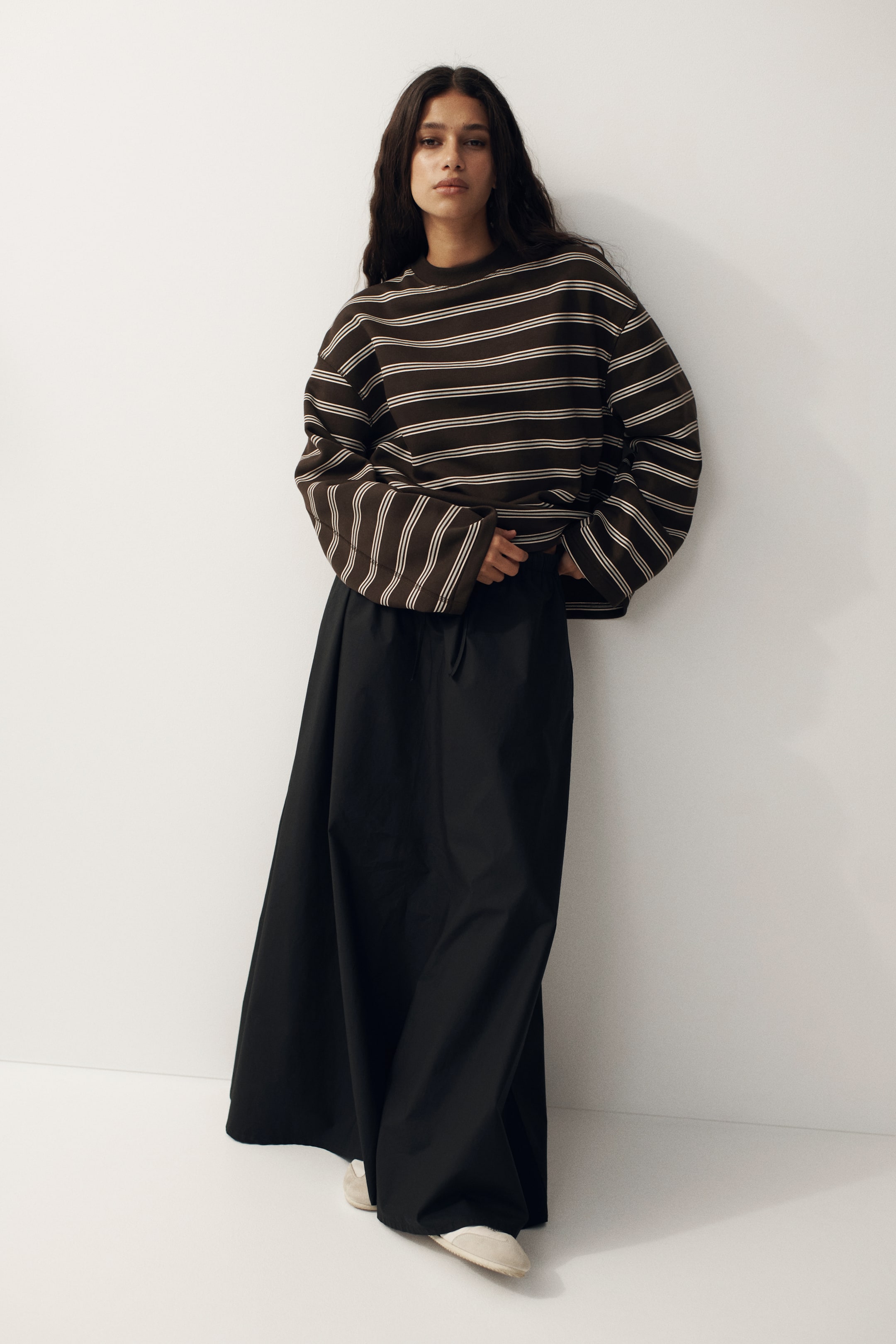 Vezi imaginea mai mare: Un hanorac maro închis, cu dungi subțiri, bej deschis, dispuse orizontal, un guler rotund și o croială supradimensionată, este purtat. Este combinat cu o fustă neagră, cu croială largă și lungime completă, care prezintă un detaliu vizibil de șnur în față.