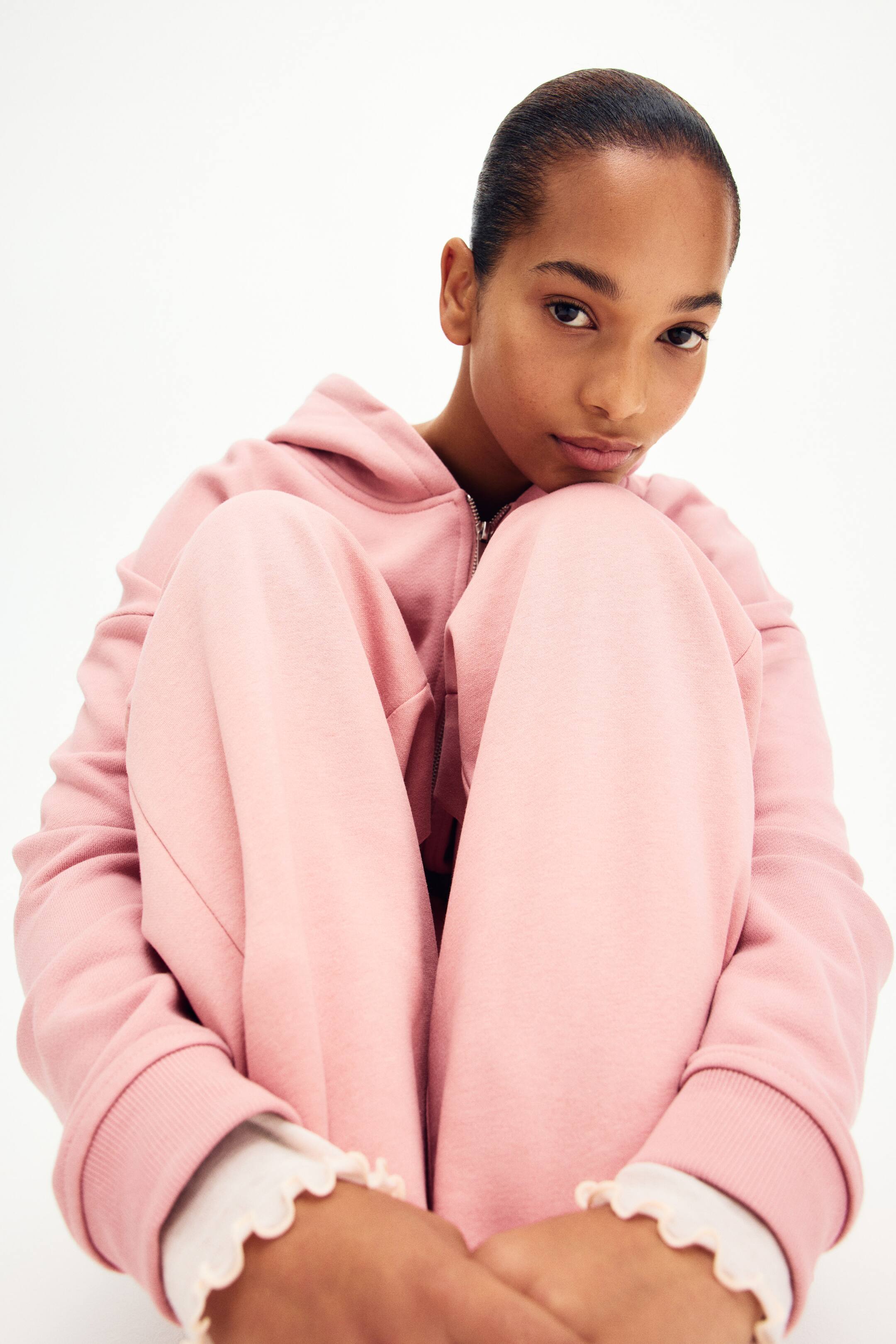 View larger image: A pink tracksuit is being worn, comprising a zippered hoodie and matching joggers. The hoodie showcases ribbed cuffs, from which a white long-sleeved top with a ruffled hem is peeping out. The joggers exhibit a relaxed fit, gathered around the wearer's bent knees.