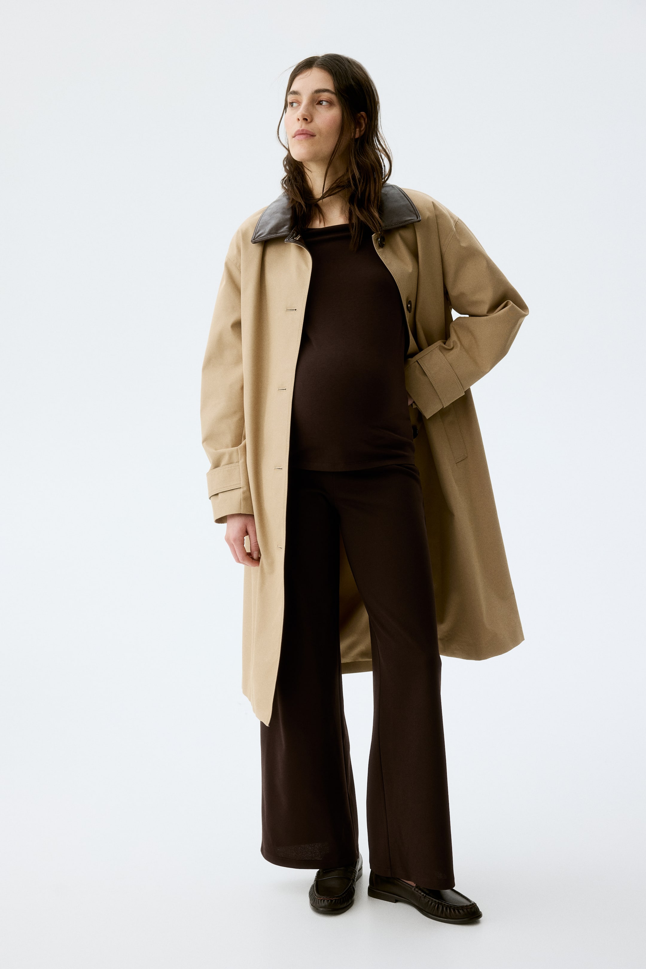 拡大画像を表示: ベージュのトレンチコートがわずかに角度をつけて紹介されています。特徴的な濃い茶色の襟が際立ちます。コートは開いた状態で着用されており、下には濃い茶色のトップスとそれにマッチしたワイドレッグパンツが見えます。