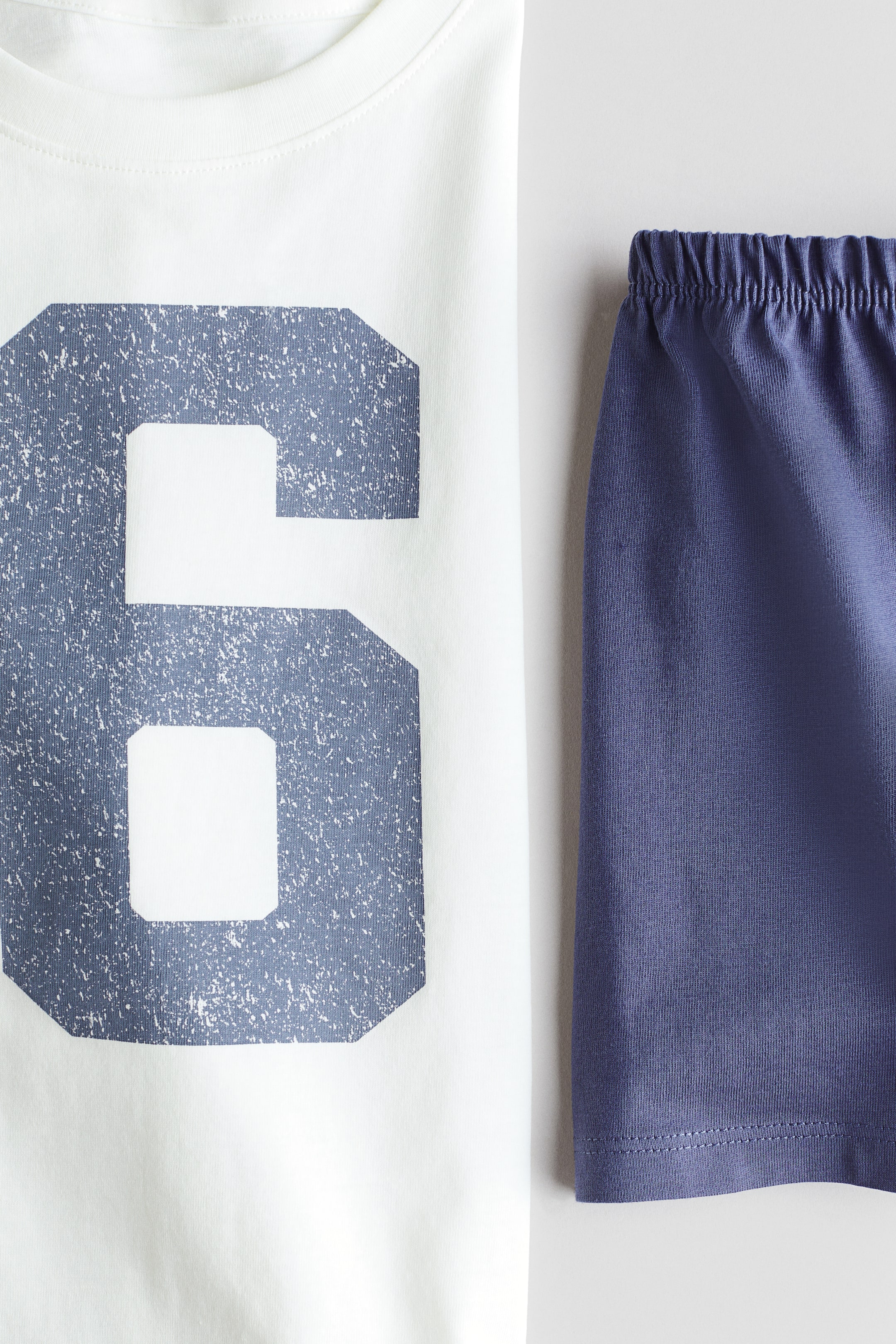 Visualizza l'immagine ingrandita: Un top pigiama in jersey di cotone bianco presenta uno scollo rotondo e una grafica con il numero '6' blu sbiadito sul davanti, abbinato a pantaloncini pigiama in jersey di cotone blu scuro con un elastico in vita arricciato.