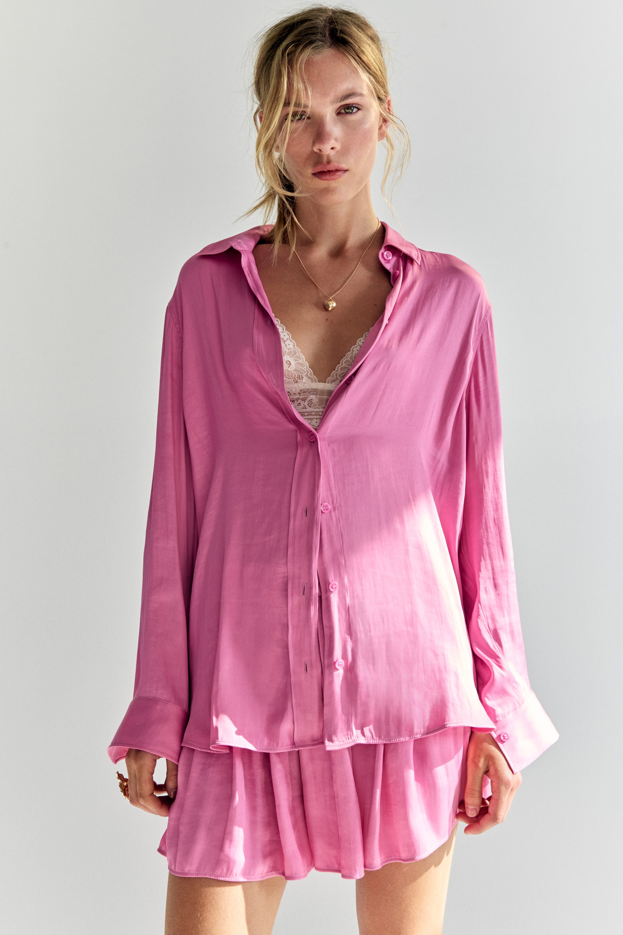 Grotere afbeelding bekijken: Een roze glanzende blouse met knoopsluiting en een bijpassend geplooid mini-rokje worden getoond. De oversized blouse, met zijn puntige kraag, lange mouwen met manchetten en knoopsluiting aan de voorkant, wordt open gedragen over een witte kanten bralette.
