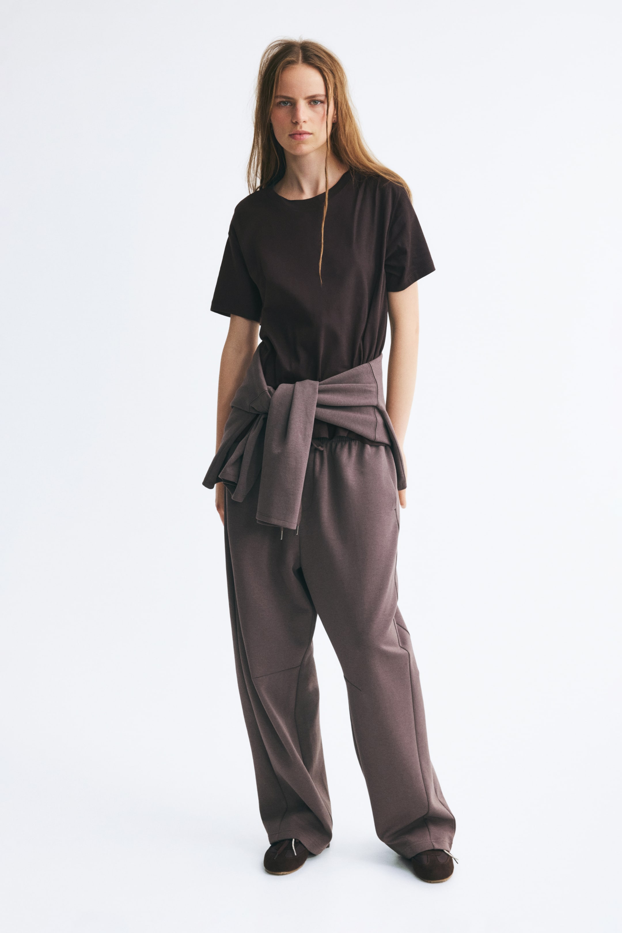 View larger image: The model is wearing a dark brown, straight-cut cotton jersey tee featuring a rib-trimmed crew neckline and softly dropped shoulders. The tee is tucked into loose-fitting, wide-leg sweatpants, with a matching sweatshirt tied around the waist.