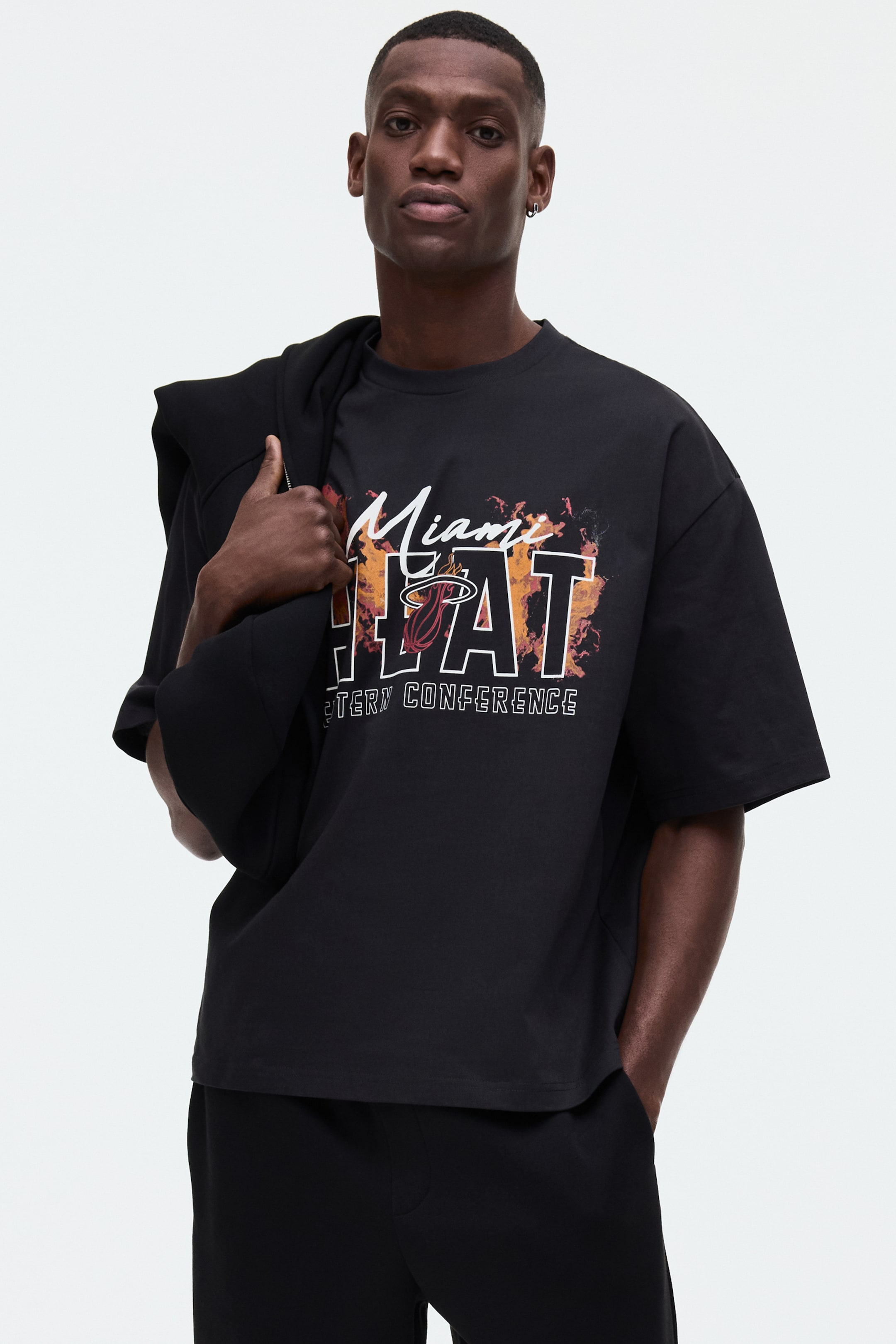 Visa större bild: En svart t-shirt med ett stort grafiskt tryck på framsidan, som visar "Miami" i vit skrivstil ovanför "HEAT" i fetstil med ett orange och rött flammmönster. "Miami Heat"-logotypen syns inom "H" i "HEAT", och "EASTERN CONFERENCE" är tryckt nedanför. T-shirten har korta ärmar och en rund halsringning, och den har en avslappnad passform.