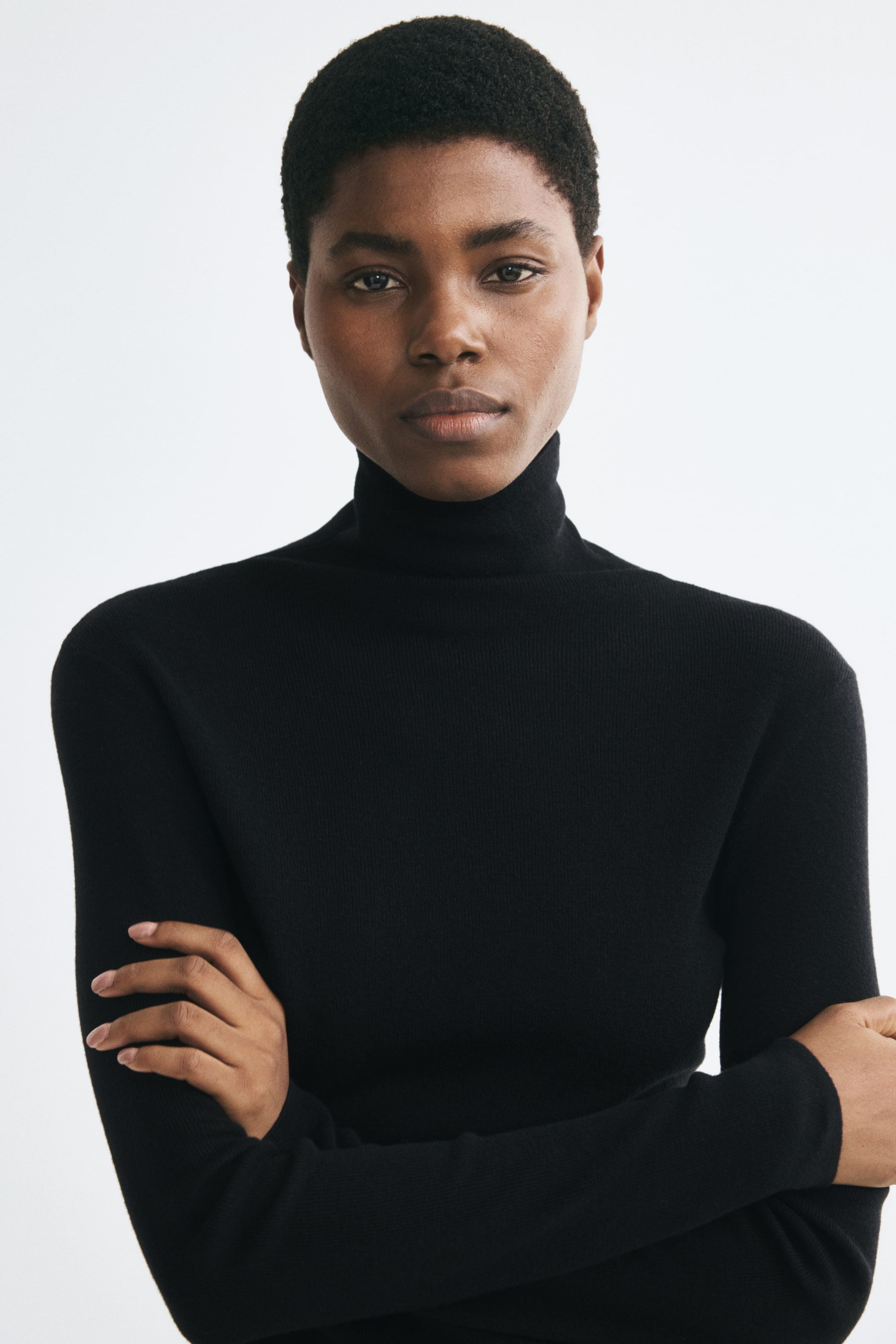 View larger image: A black, fitted roll-neck sweater in a fine knit is being worn by the model. The sweater features a high roll-neck collar and long sleeves, with the model posing with crossed arms.
