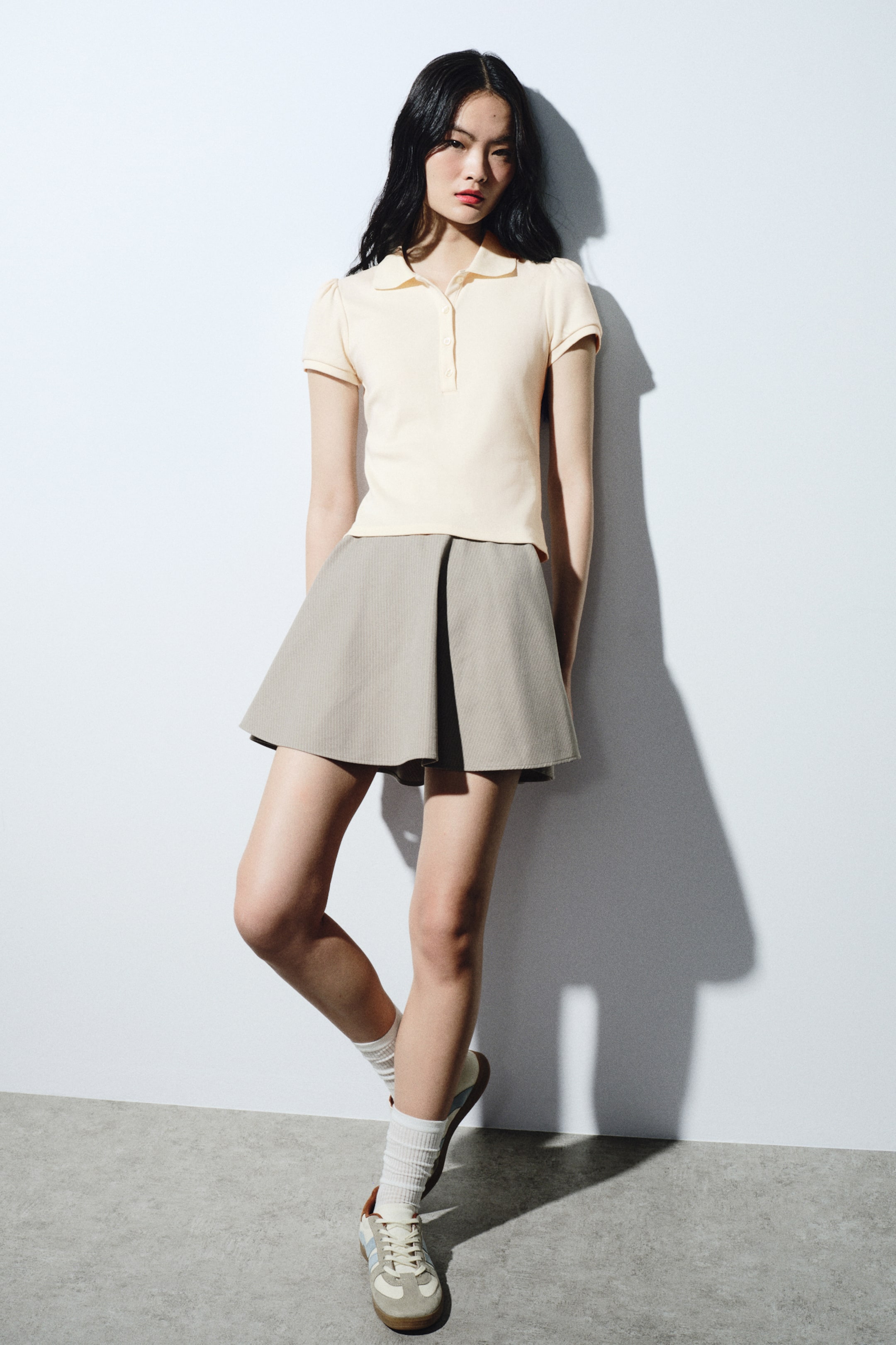拡大画像を表示: 淡いイエローのポロシャツは、先の尖った襟、3つボタンの前立て、リブカフス付きのショートパフスリーブを備えています。淡いグレーのミニスカートは、さりげない縦ストライプ柄とフレアのAラインシルエットが特徴です。白いリブのクルーソックスがガムソールのベージュ×ライトブルーのロープロファイルスニーカーにだぶっと被さっています。