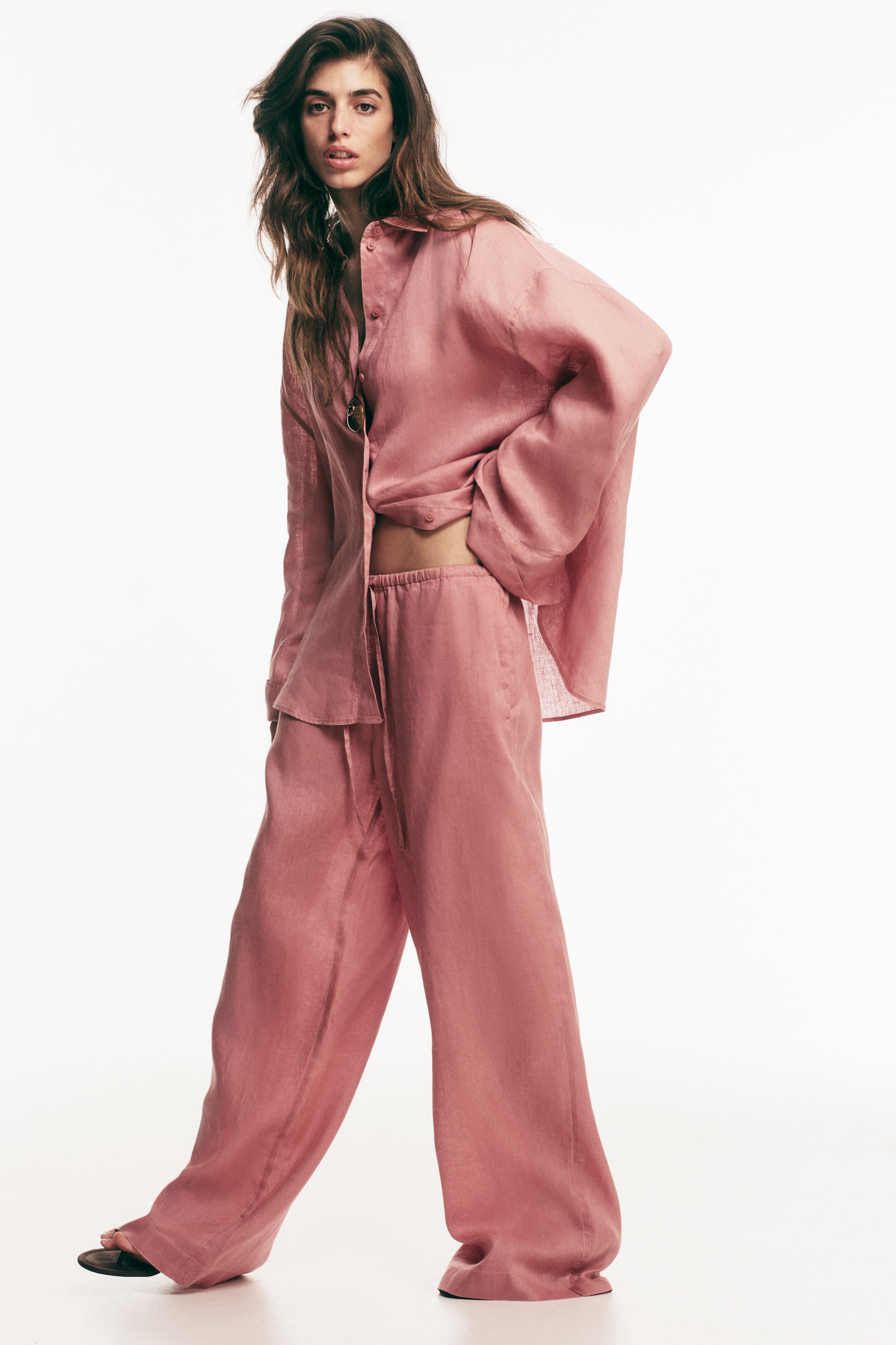 View larger image: Loose-fit pink trousers in breezy linen with an elasticated, drawstring waist and wide legs are worn by the model. The model rests one hand on the waistband, also donning a matching loose-fit linen shirt.