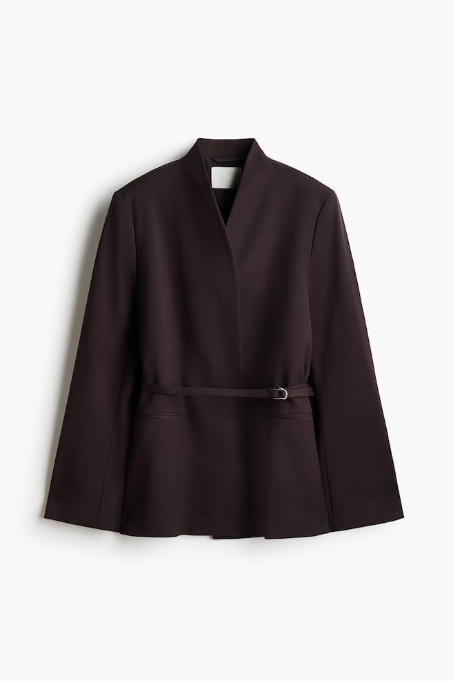 An oversized dark brown blazer is shown from the front, featuring structured shoulders, a stand collar, and a wrap-front closure secured by a narrow matching belt at the waist. The design includes subtle jetted front pockets and long, wide sleeves. - 1