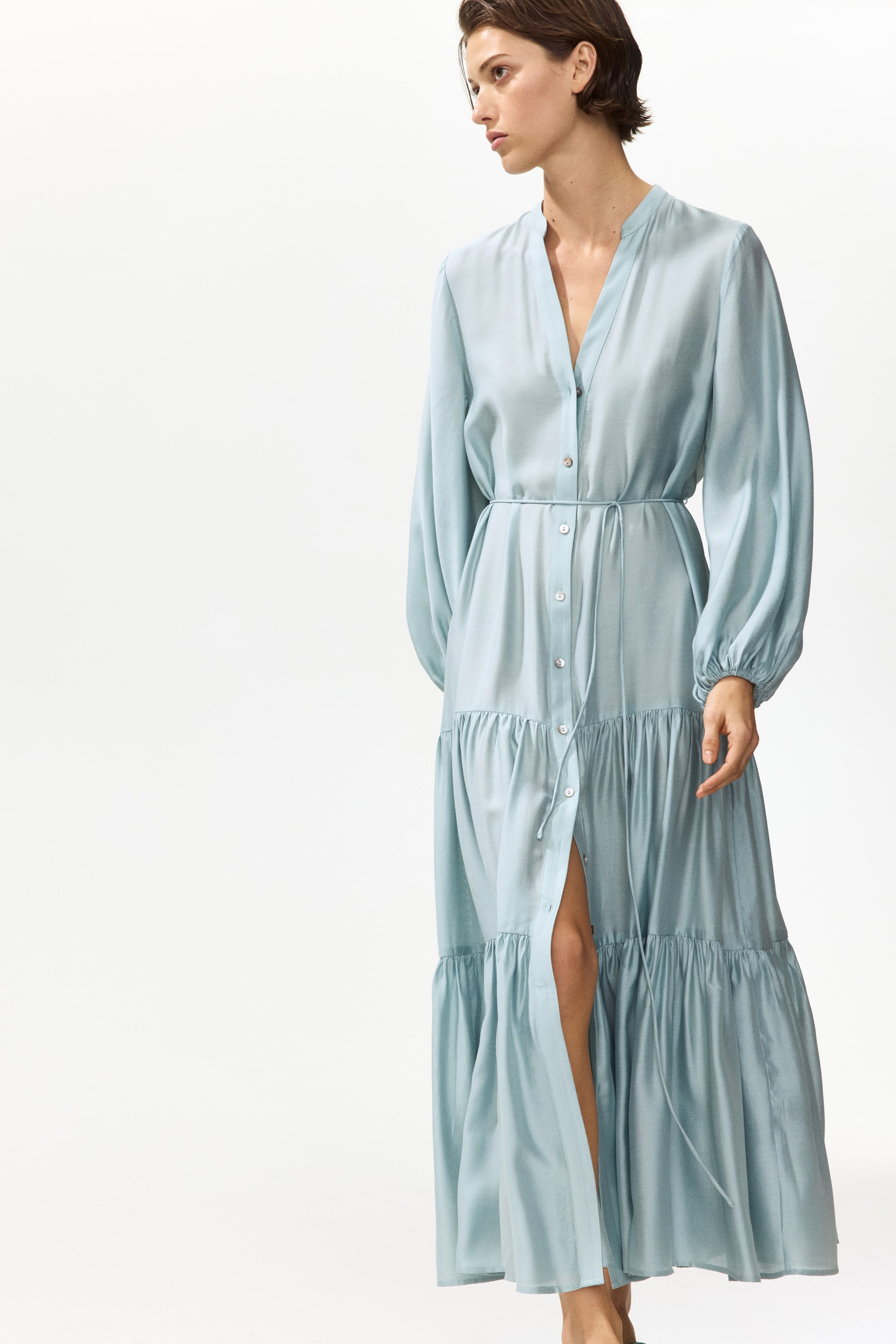 Visa större bild: En ankellång ljusblå klänning med en lätt glans bärs av modellen och har en ståkrage, V-formad öppning, knappar längs framsidan, 3/4-långa ballongärmar med elastiska muddar, ett smalt knytband i midjan och en skiktad A-linjeformad kjol. Modellen poserar och visar ett ben genom den delvis uppknäppta framsidan.