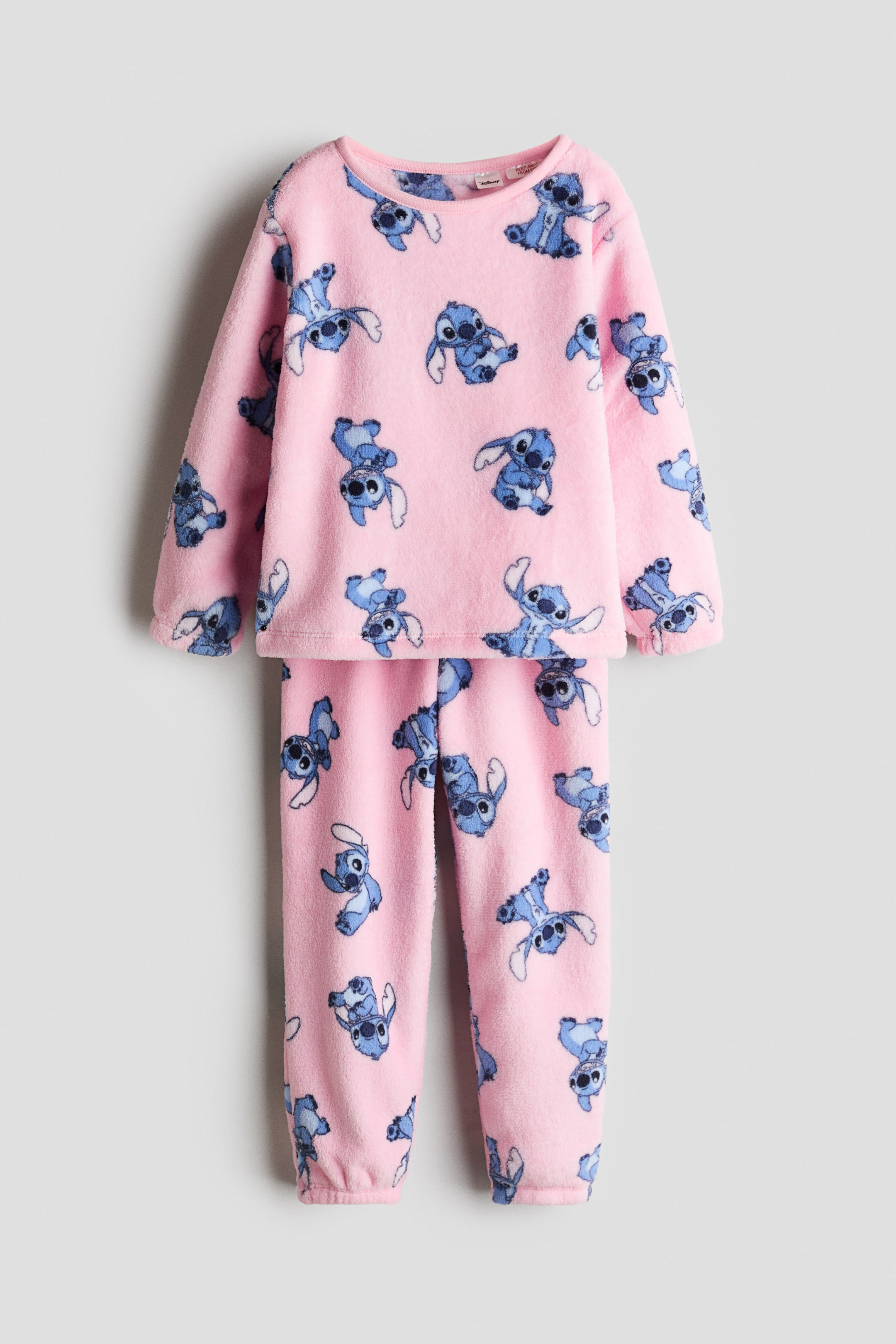 Näytä suurempi kuva: Pyjamapari on esitetty edestäpäin, ja siinä on vaaleanpunainen, pehmeä veluurikangas ja sininen hahmoprintti. Yläosassa on pyöreä pääntie, pitkät hihat ja joustavat hihansuut, kun taas yhteensopivissa alaosissa on joustava vyötärö ja resorit lahkeensuissa.