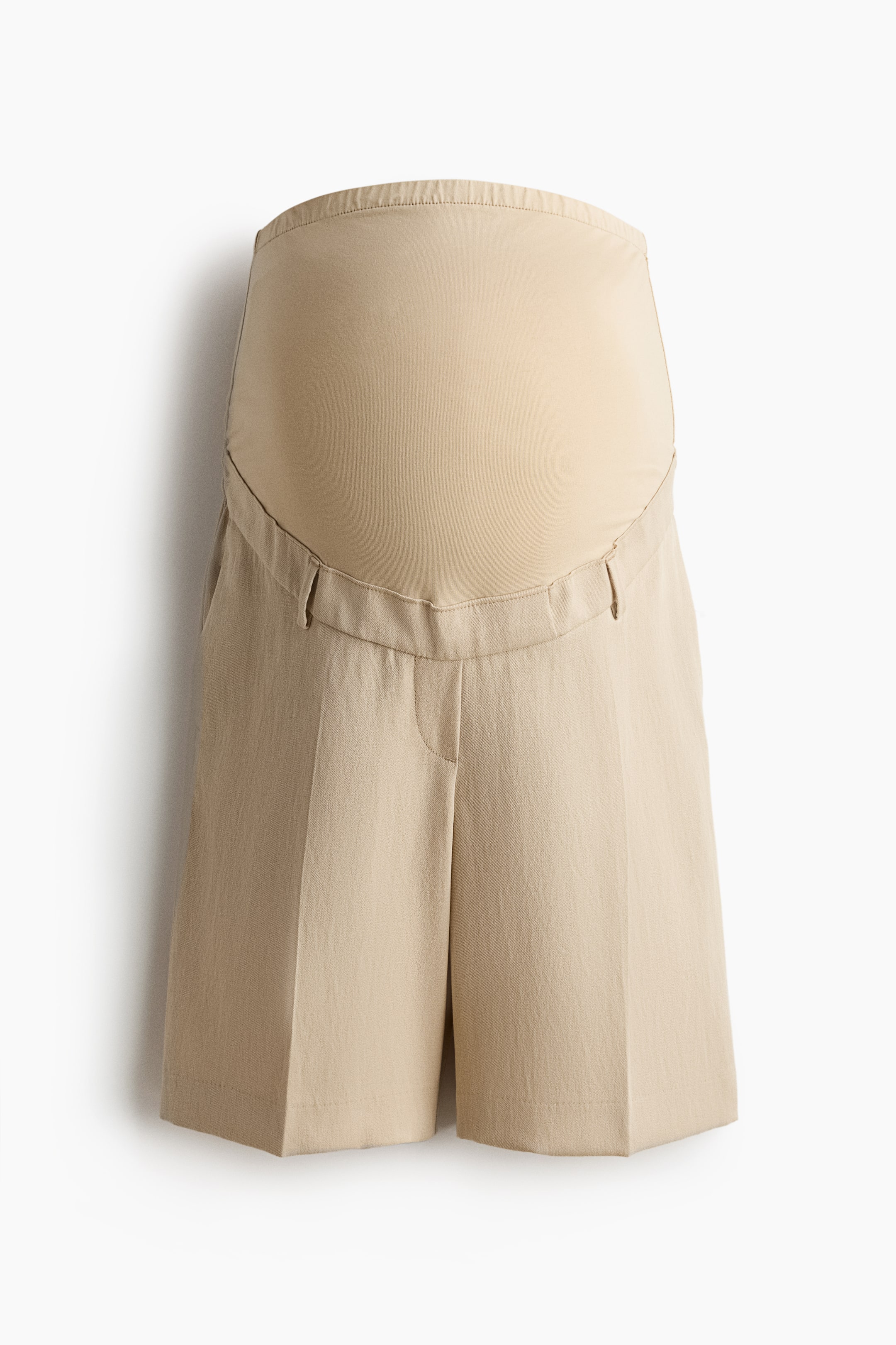 Visualizza l'immagine ingrandita: I pantaloncini premaman sono presentati in vista frontale, con un pannello per il ventre in beige chiaro, liscio ed elastico, caratterizzato da un bordo superiore finemente a coste. La sezione principale dei pantaloncini è realizzata in un tessuto beige leggermente più scuro e strutturato, progettata con un fronte piatto e piccoli passanti per cintura, terminando sopra il ginocchio.