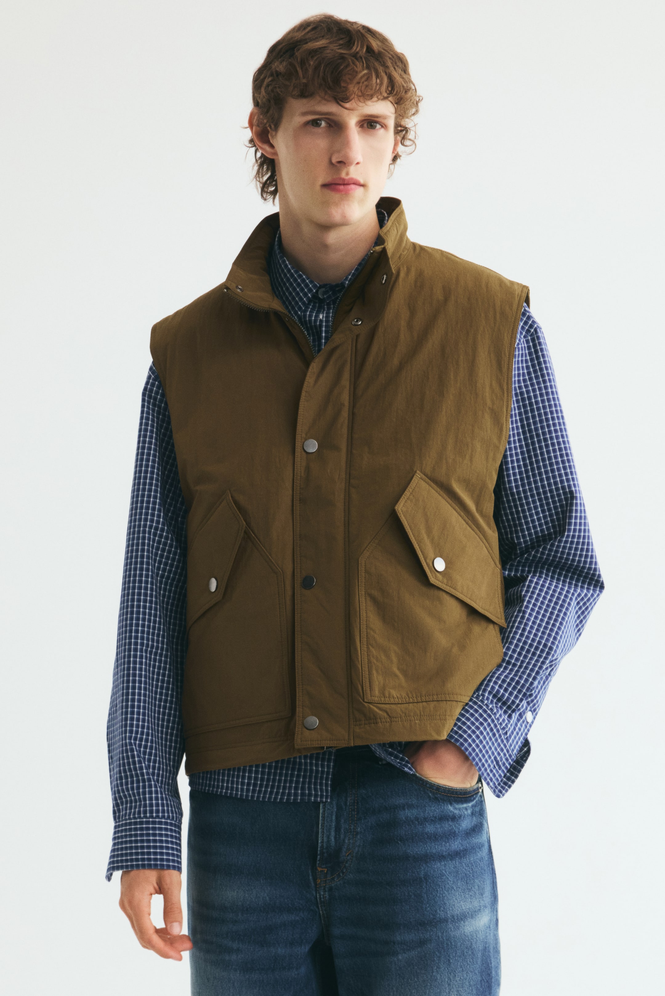 Grotere afbeelding bekijken: Een bruine, licht gewatteerde bodywarmer met opstaande kraag, een sluiting met drukknopen aan de voorkant en grote opgestikte zakken met kleppen en drukknoopsluiting wordt gedragen door het model. De bodywarmer is gelaagd over een blauw-wit geruit hemd met lange mouwen en een blauwe jeans.