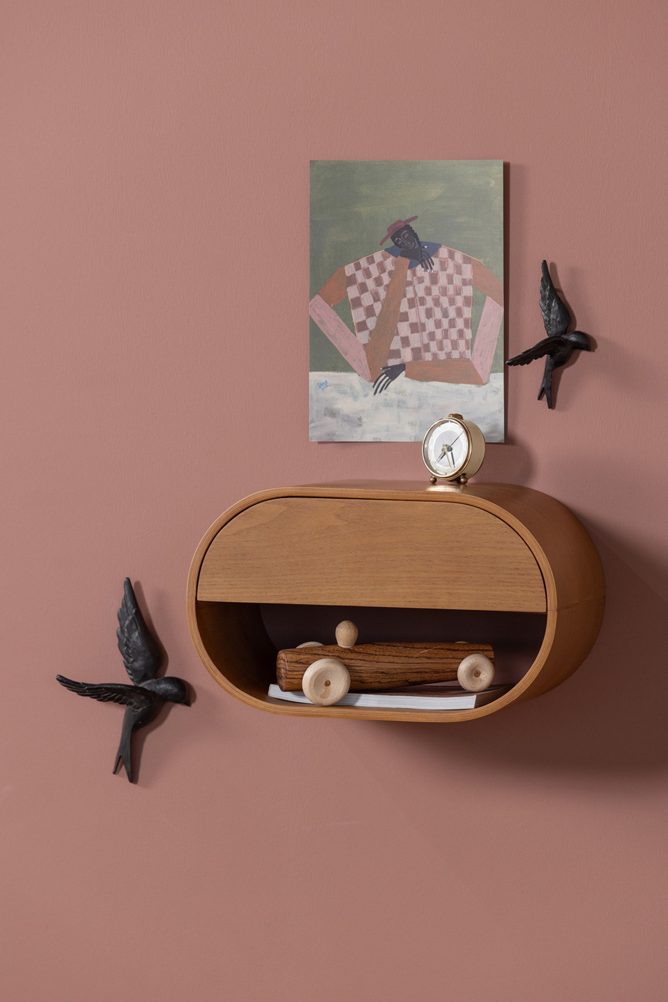 Grotere afbeelding bekijken: Een ovale, aan de muur gemonteerde houten plank met een lichte houtafwerking, heeft een gebogen lade in het bovenste gedeelte en een open opbergvak eronder. Een kleine wekker rust op het bovenoppervlak en een houten speelgoedauto staat in het onderste vak.