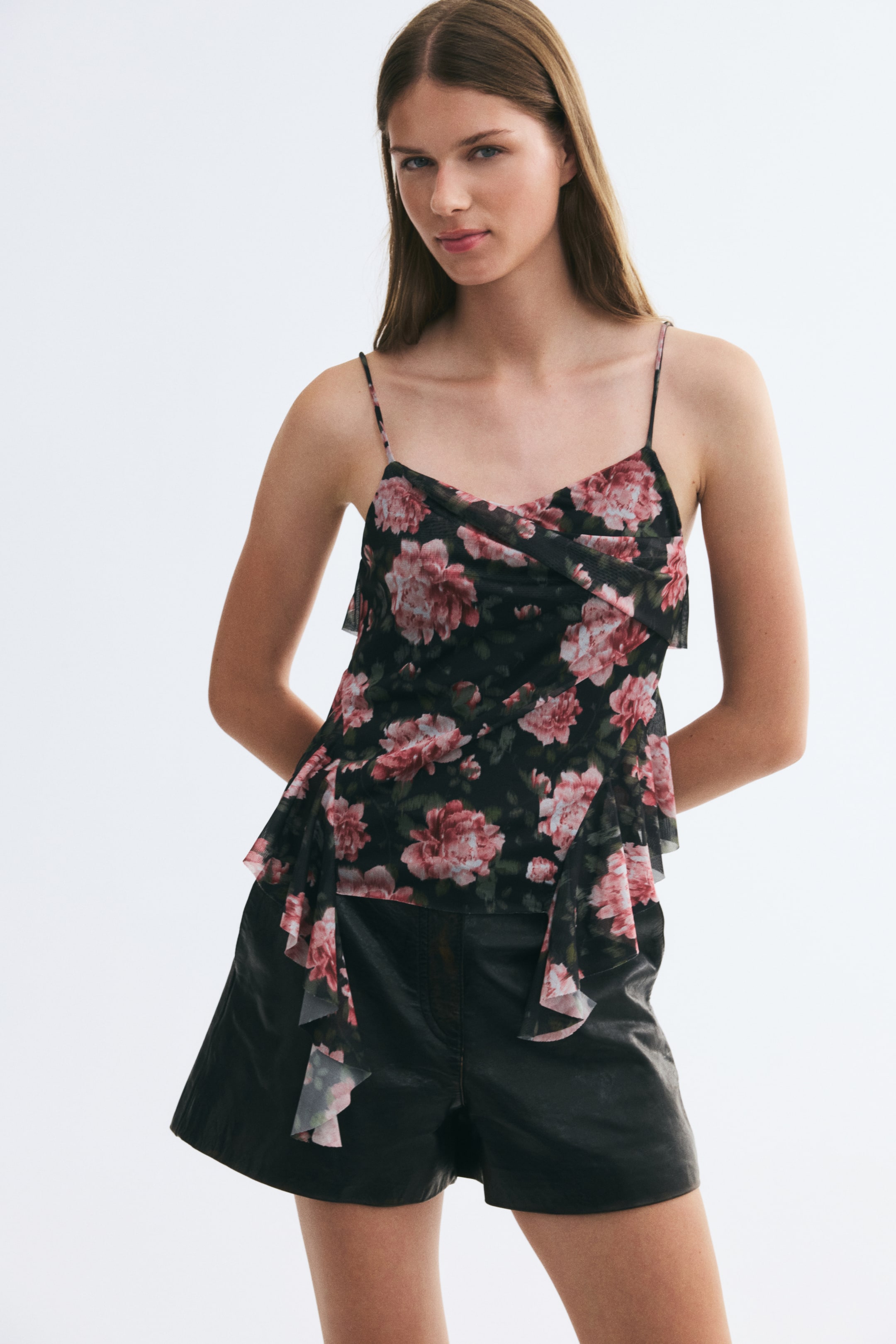 View larger image: A sheer mesh camisole in a dark floral print is worn by the model, paired with black shorts. The top features spaghetti straps, an asymmetrical foldover front, and draped, flounced panels cascading down the sides.