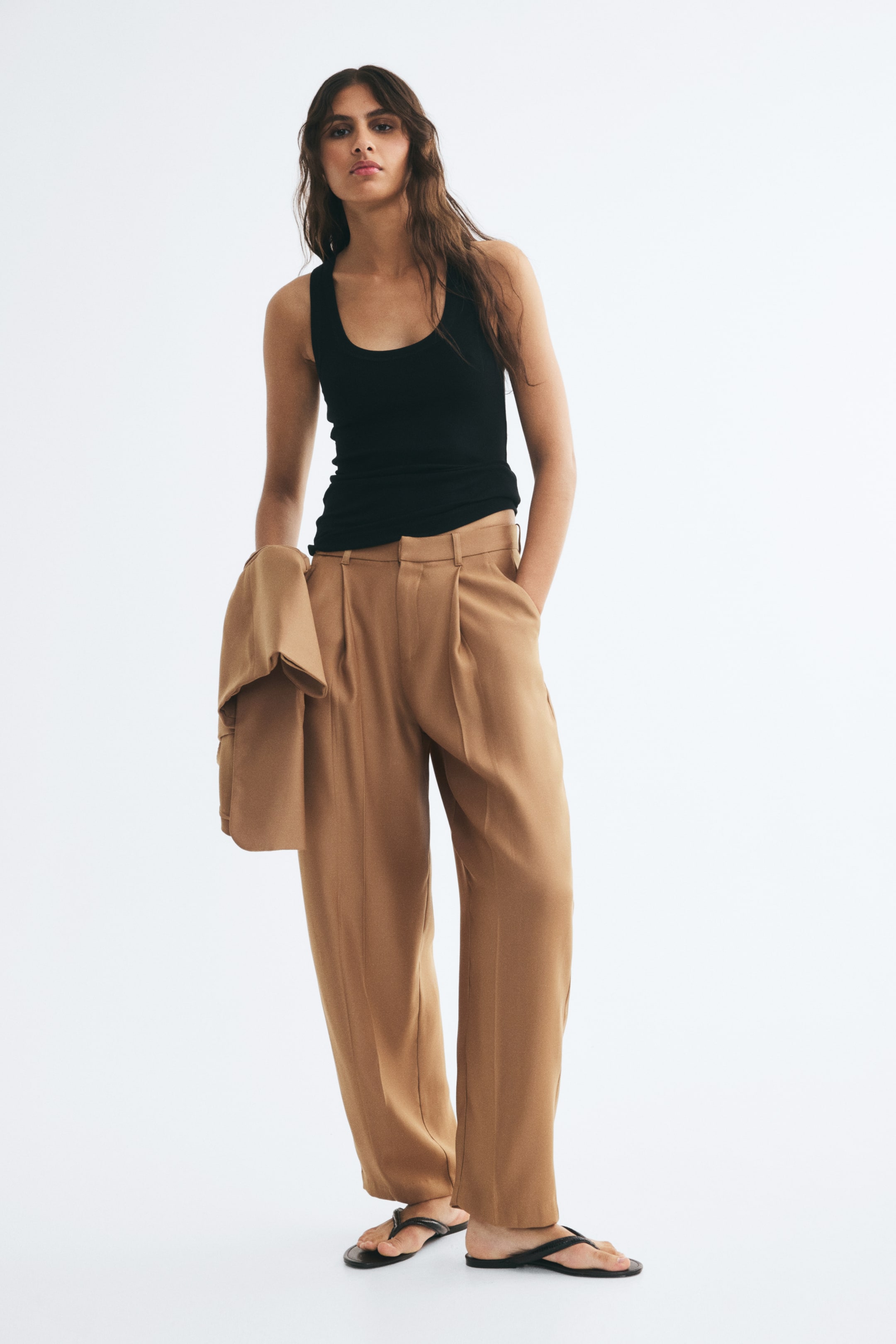 View larger image: The model is wearing wide-leg, pleated camel-colored trousers with one hand resting in a side pocket. A matching camel blazer is draped over the model’s arm, paired with a black scoop-neck tank top.