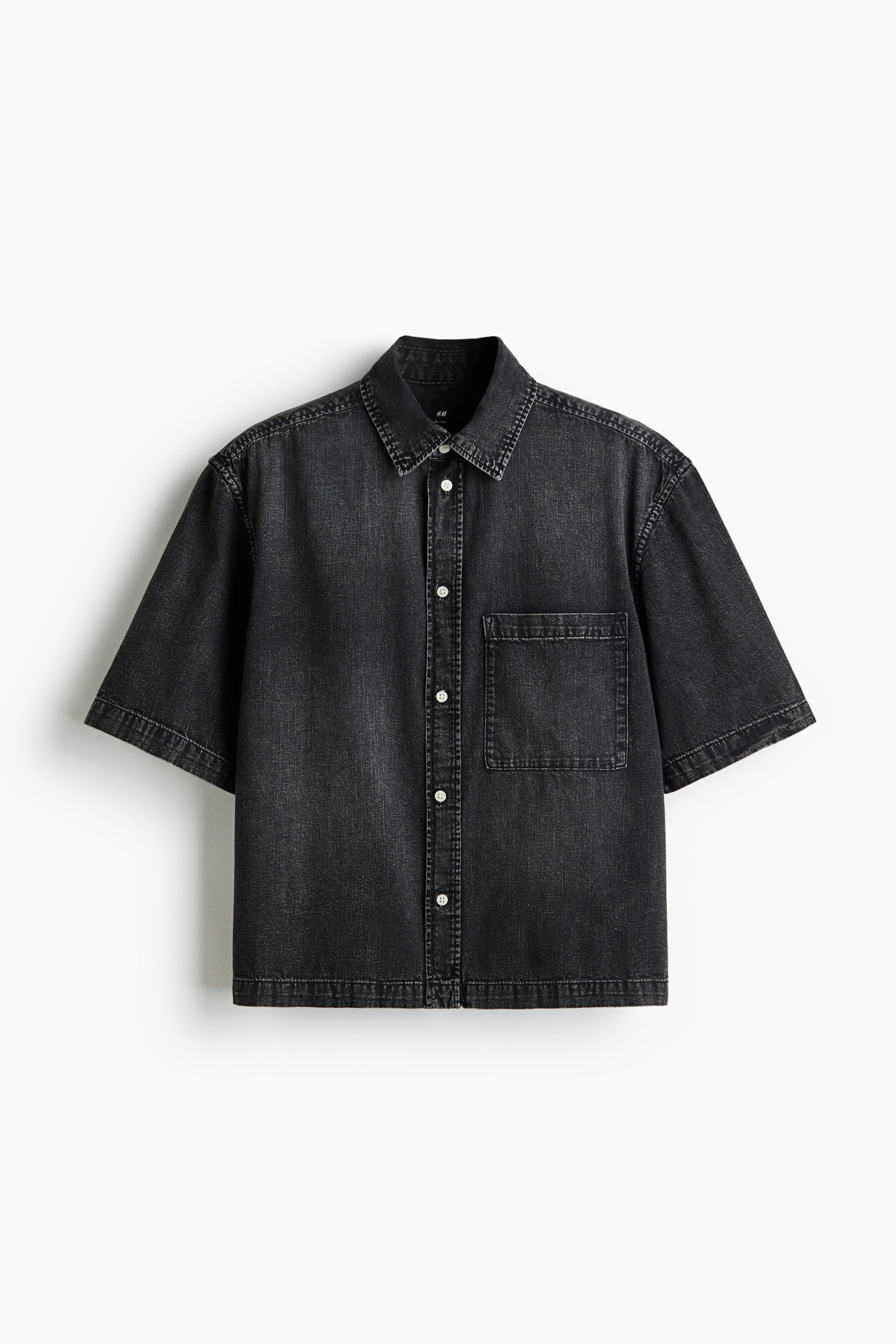 View larger image: A washed black denim short-sleeve shirt is shown from the front, featuring a pointed collar, full button-front closure with light-colored buttons, and a single patch pocket on the left chest. It has wide short sleeves, visible contrast stitching, and a relaxed fit.